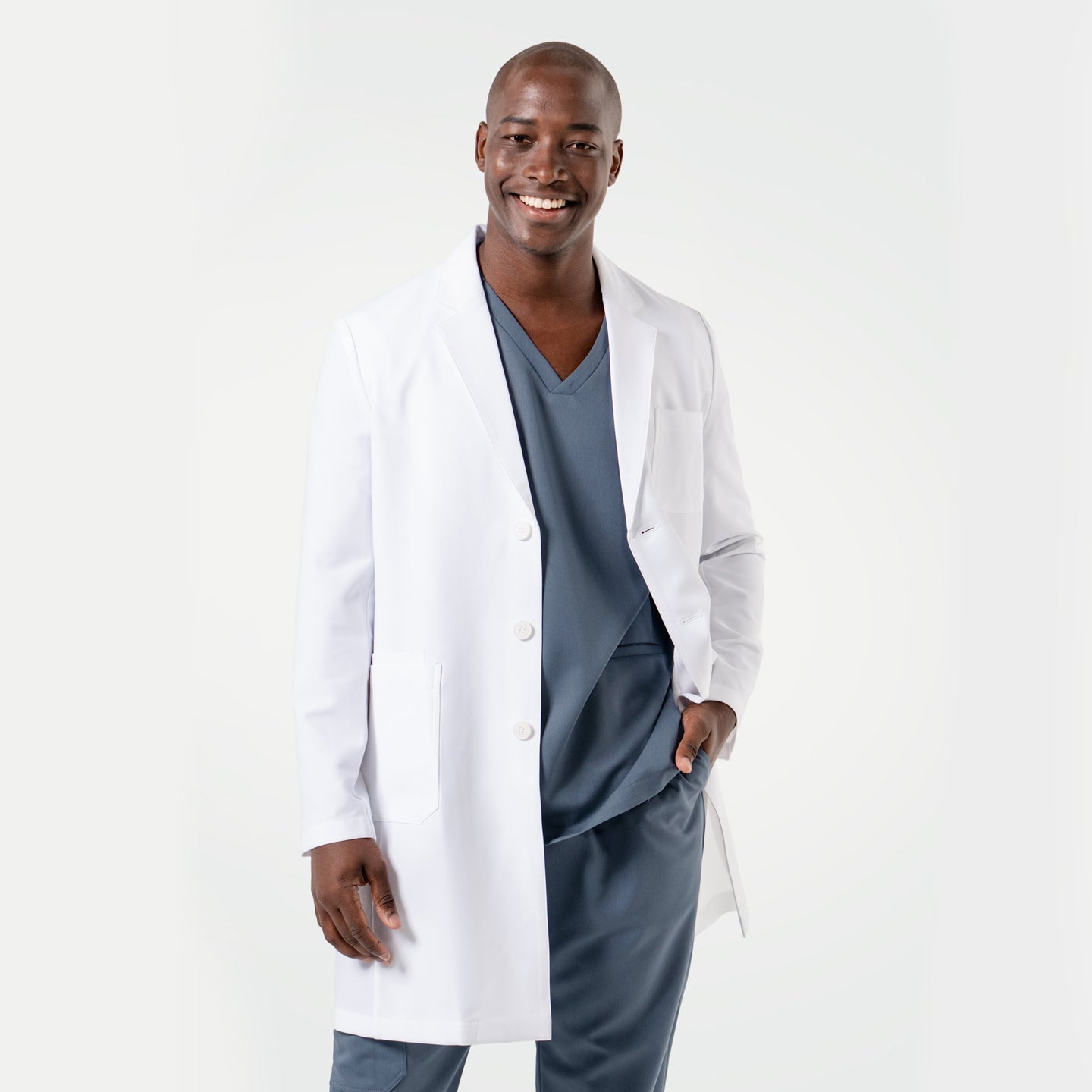 Smiling in the Men's LCM-101 Lab Coat in White over medical scrubs, one hand in the pocket, full body view