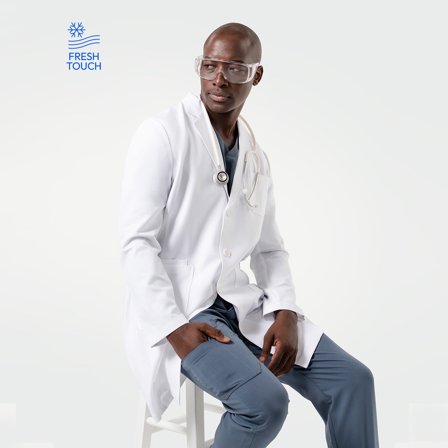 Seated in the Men's LCM-101 Lab Coat in White over medical scrubs, with a stethoscope around his neck and protective glasses, looking to the side
