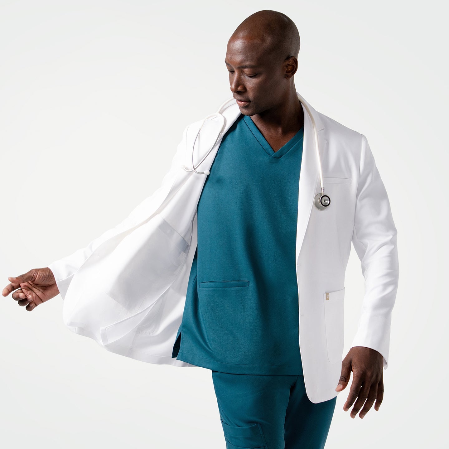 Wearing the Men's LCM-102 Lab Coat in White over medical scrubs, looking down while holding one side of the coat with a stethoscope around the neck
