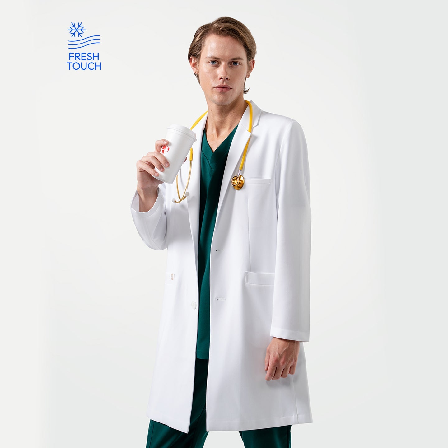 Male doctor wearing a white lab coat over green scrubs with a yellow stethoscope around his neck, holding a white takeaway coffee cup