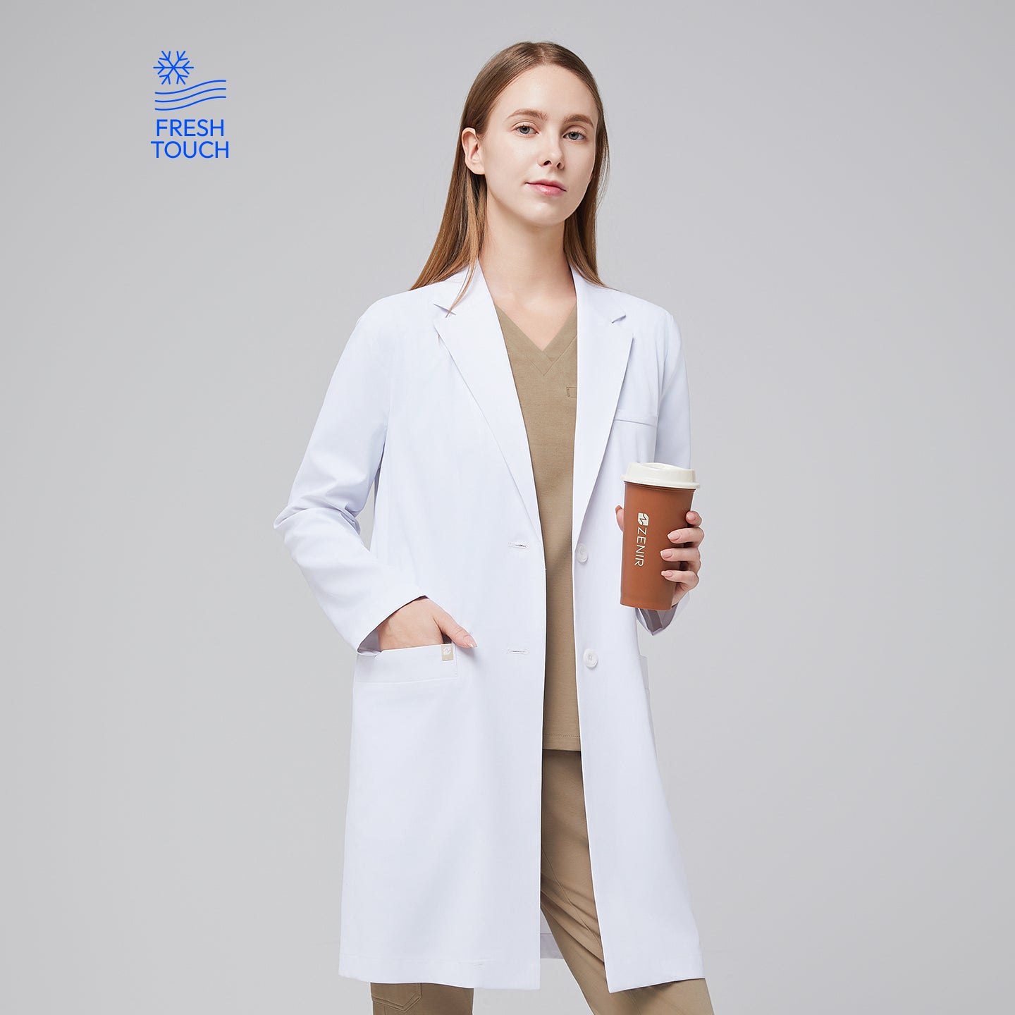 Model facing forward with a hand in her pocket, wearing a white long lab coat with chest and side pockets, with a sand beige scrub top and bottom under, holding a brown tumbler,White