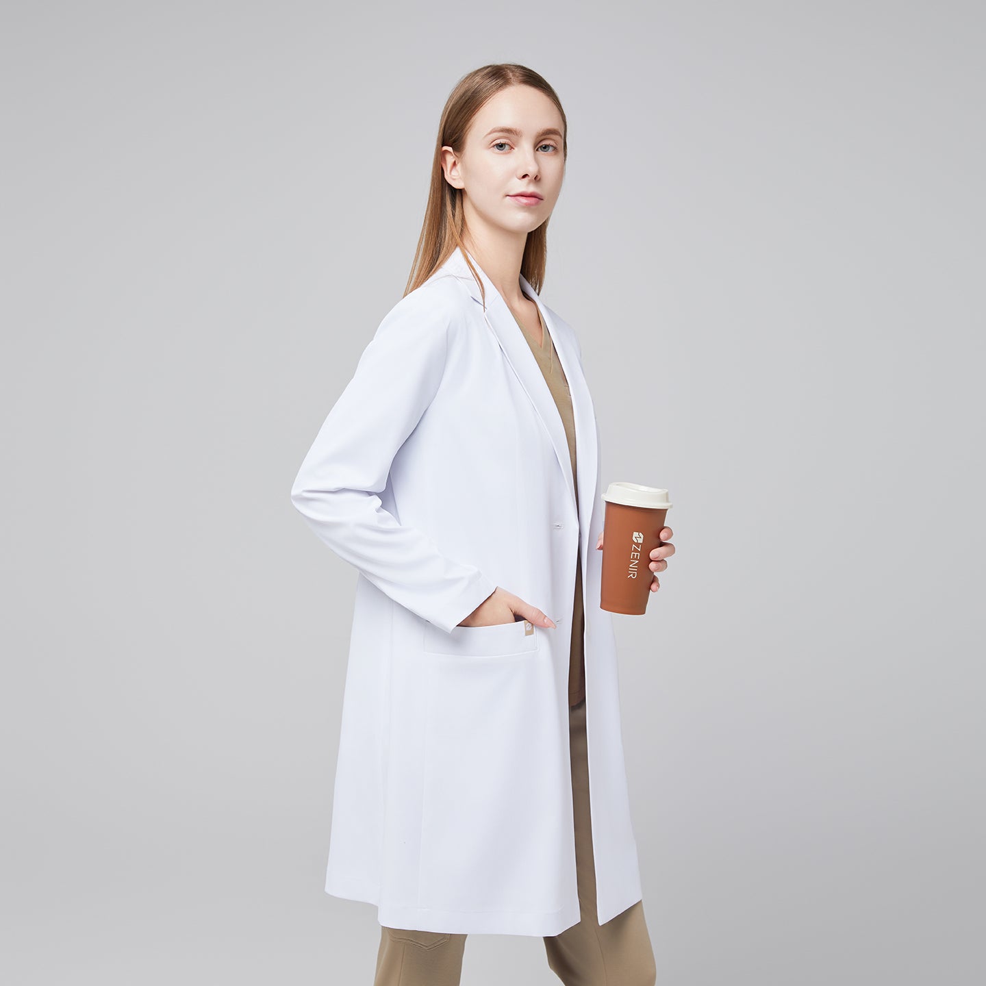 Model standing to the side with a hand in her pocket, wearing a white long lab coat with chest and side pockets, with a sand beige scrub top and bottom under, holding a brown tumbler,White