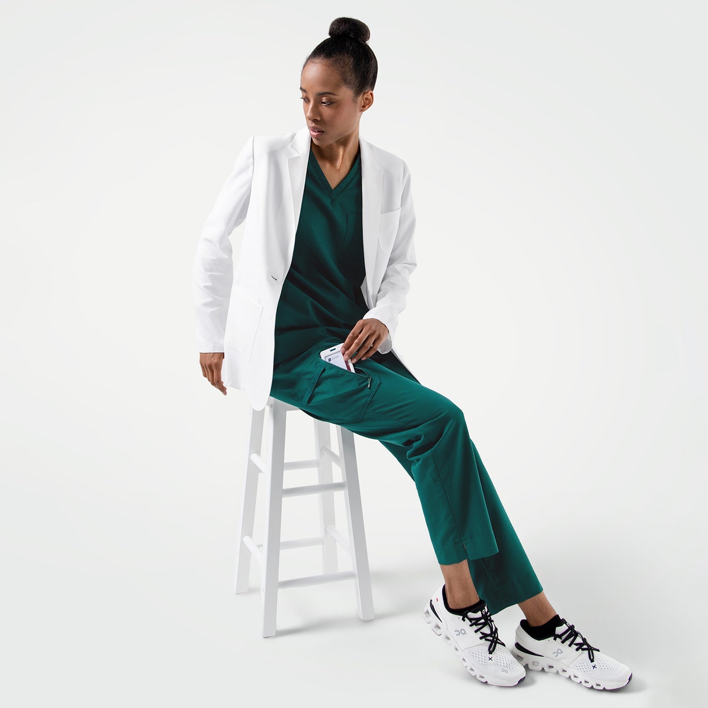 Full body shot of the model sitting on a white chair, wearing the Women's LCW-101 Lab Coat over pine green scrubs, looking slightly down while taking a phone out of her pants pocket
