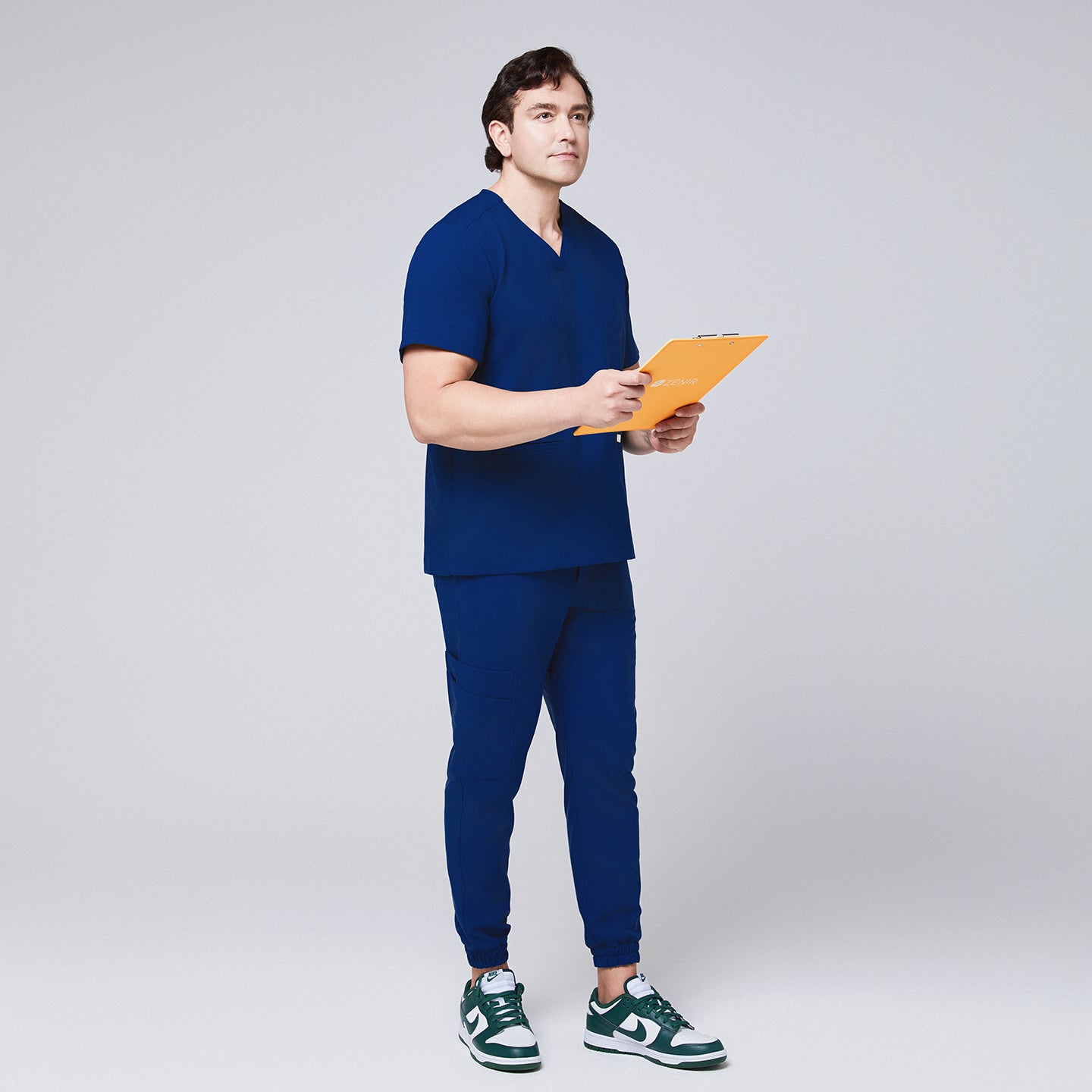 Full-body view of a man in Zenir Admiral Blue jogger scrub pants and matching top, holding a yellow clipboard, styled with green sneakers, Admiral Blue