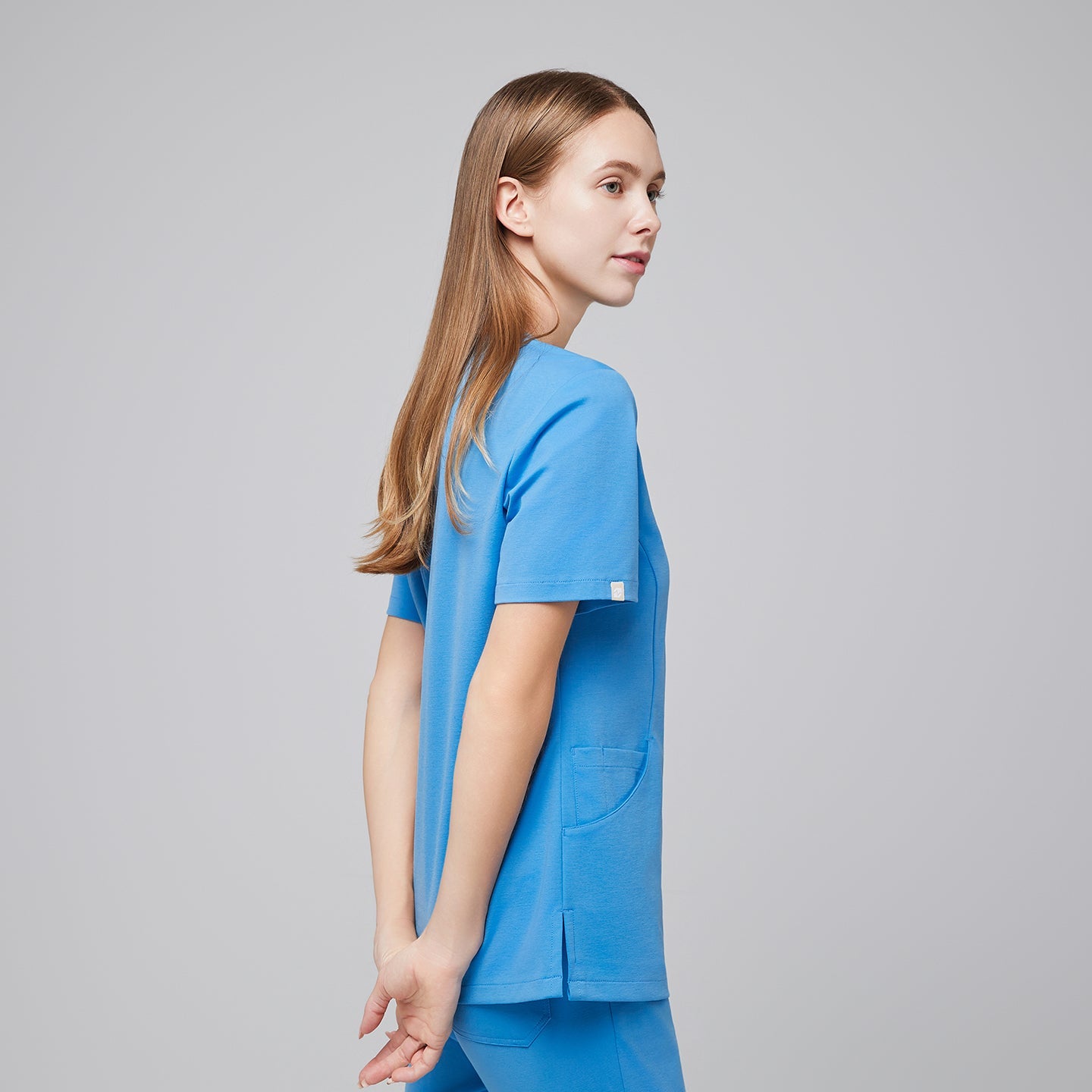 Woman in an aqua blue scrub top and pants, standing in a side pose with hands gently clasped behind her back, looking confidently to the side,Aqua Blue