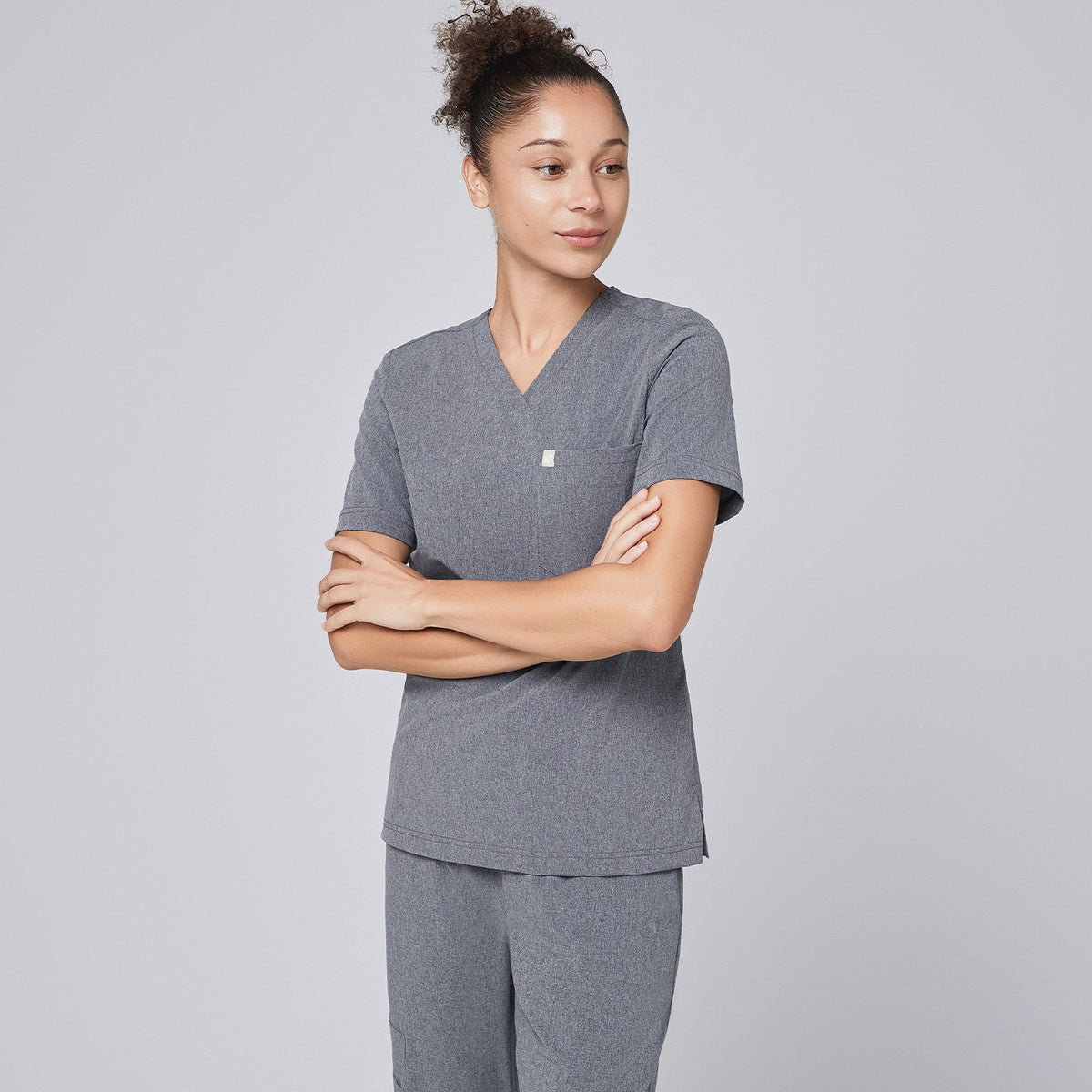 Female healthcare professional in an ash gray scrub set, standing in a rear-side pose with hands in pockets and wearing black sneakers,Ash Gray