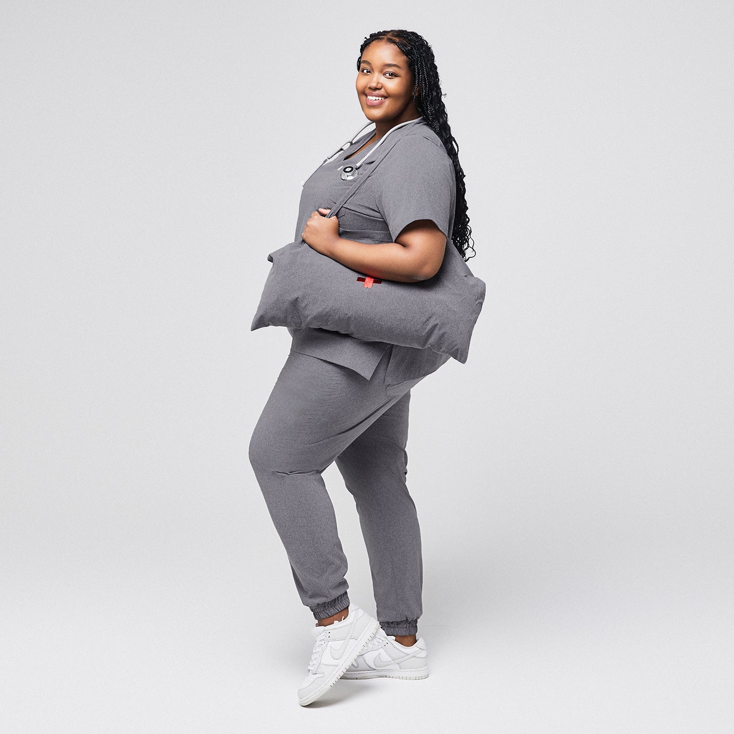 Smiling woman wearing ash gray scrub set and jogger pants, holding a matching gray tote bag with a stethoscope around her neck,Ash Gray