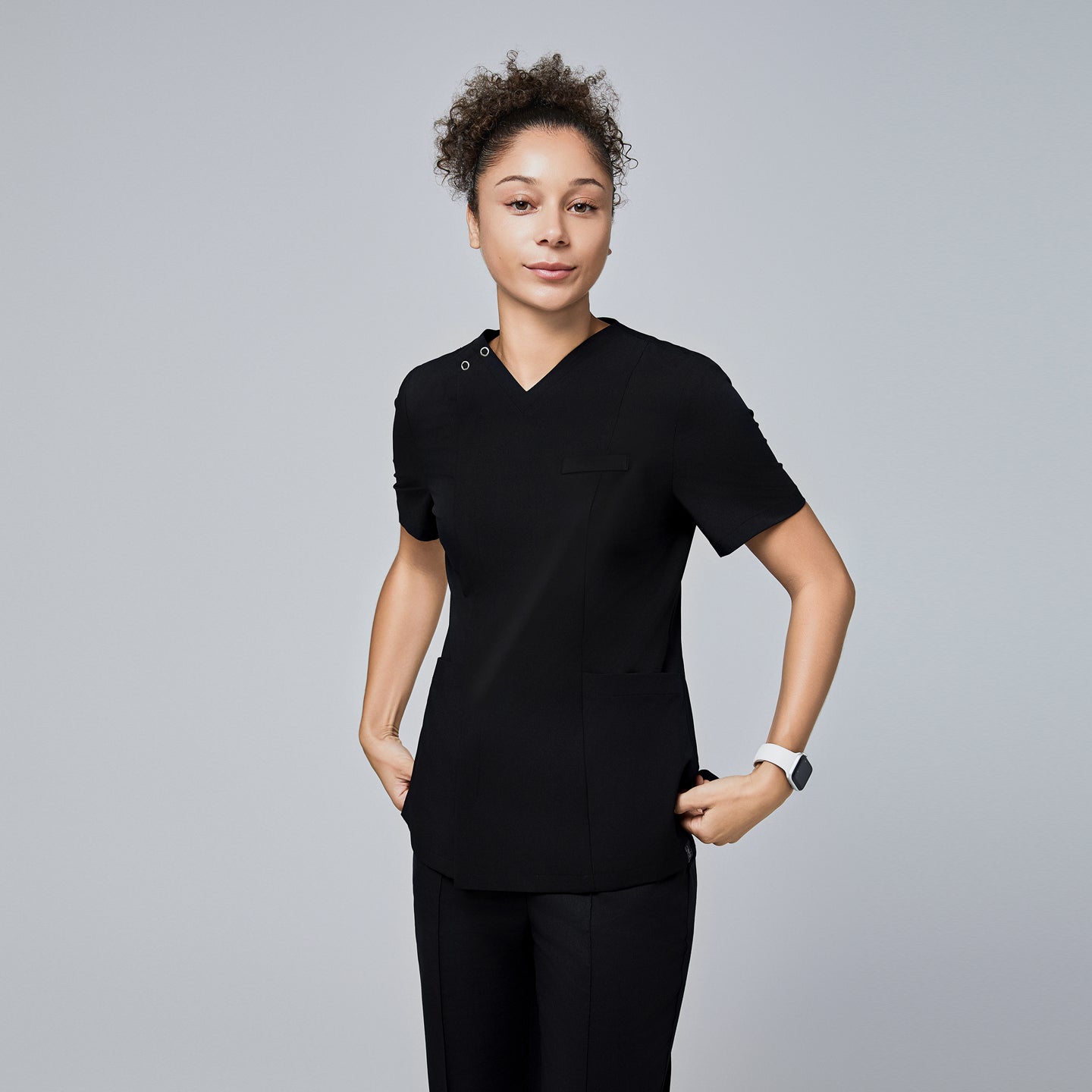 Female model wearing a black V-neck scrub top with a front zipper, short sleeves, and multiple pockets, standing with hands in pockets,Eco Black