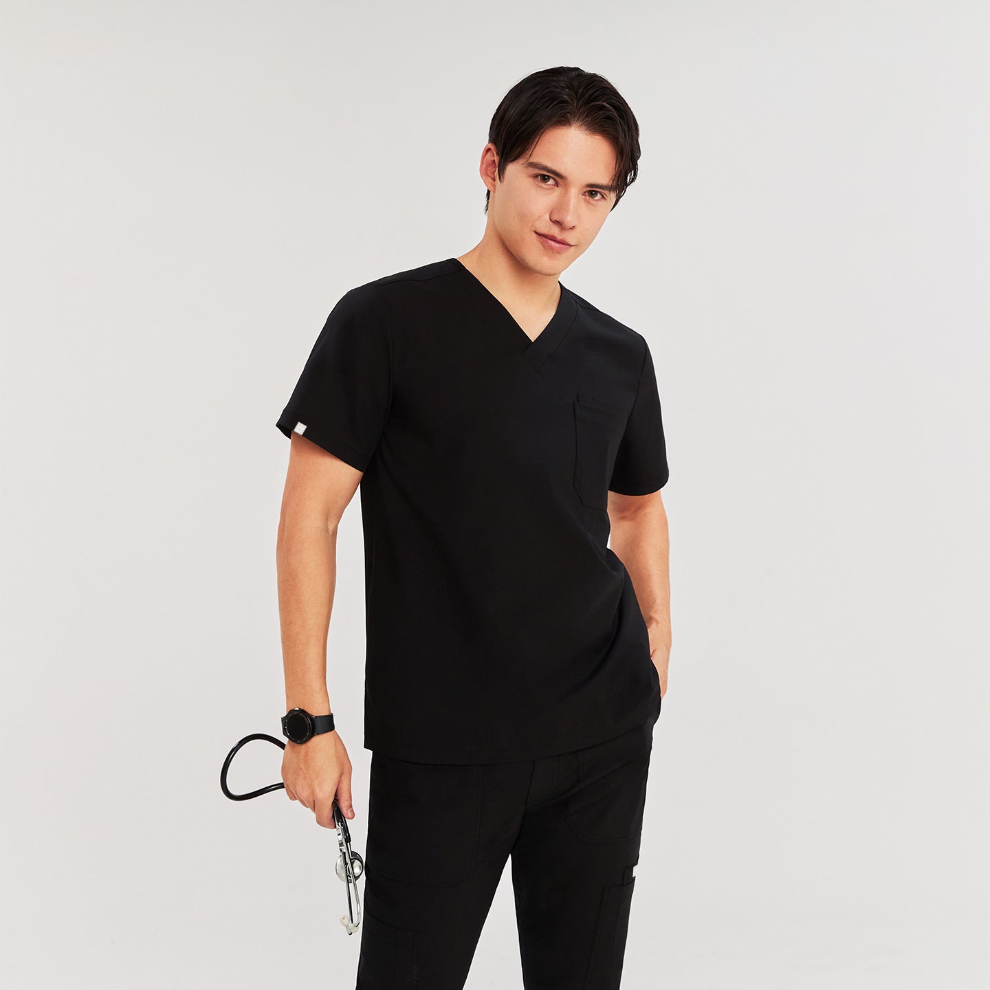 Man wearing a scrub top, holding a stethoscope, with a hand in the pocket and standing in a relaxed pose,Eco Black