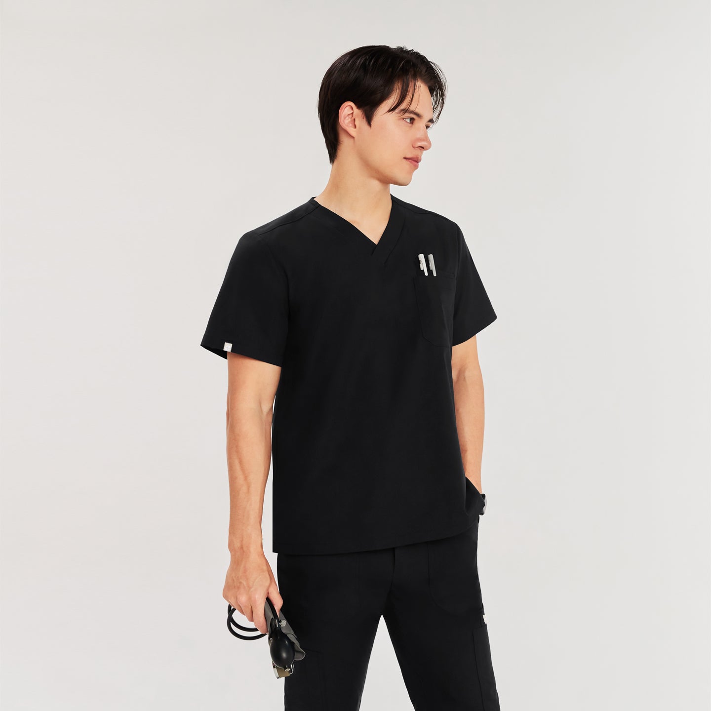 Man in black Zenir scrub top with chest pocket holding pens, standing with sphygmomanometer in hand, looking to the side,Eco Black