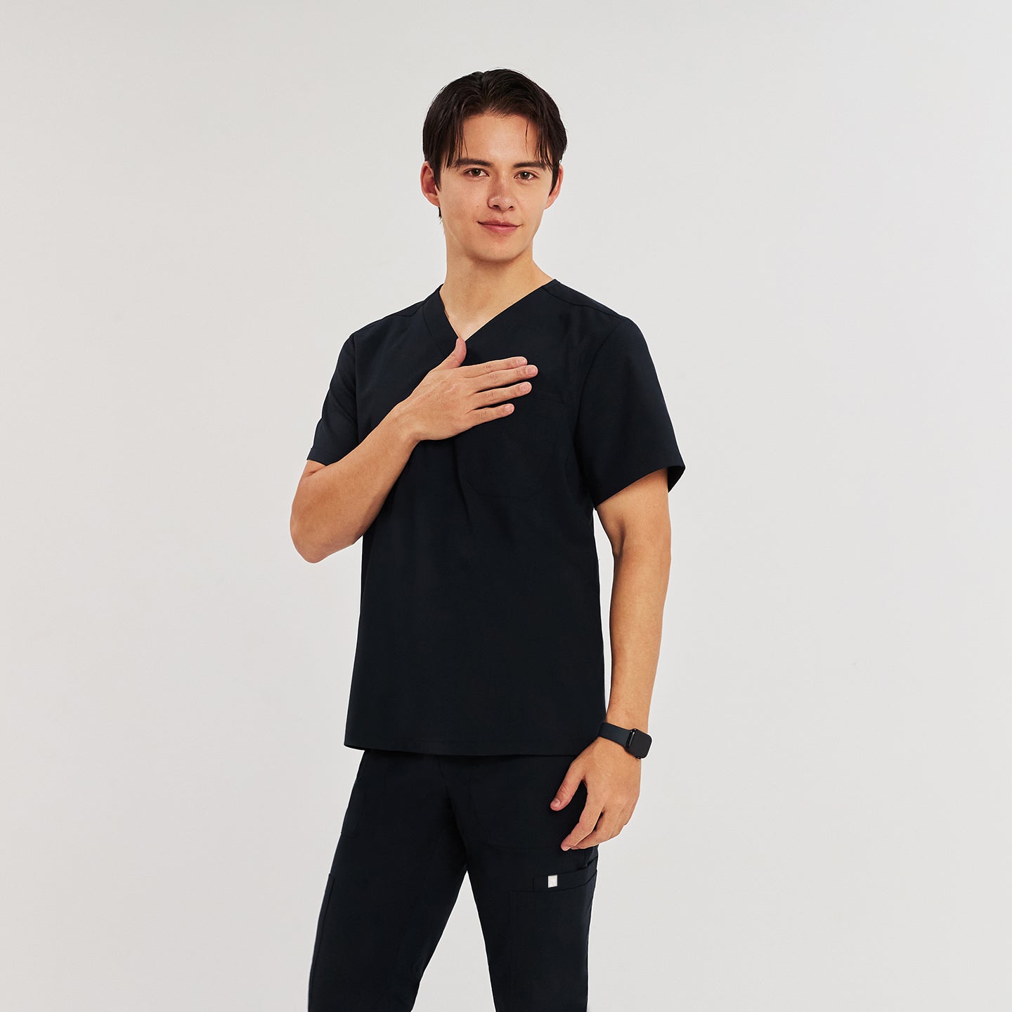 Man in black Zenir scrub top with chest pocket, standing with hand on chest, looking at the camera,Eco Black
