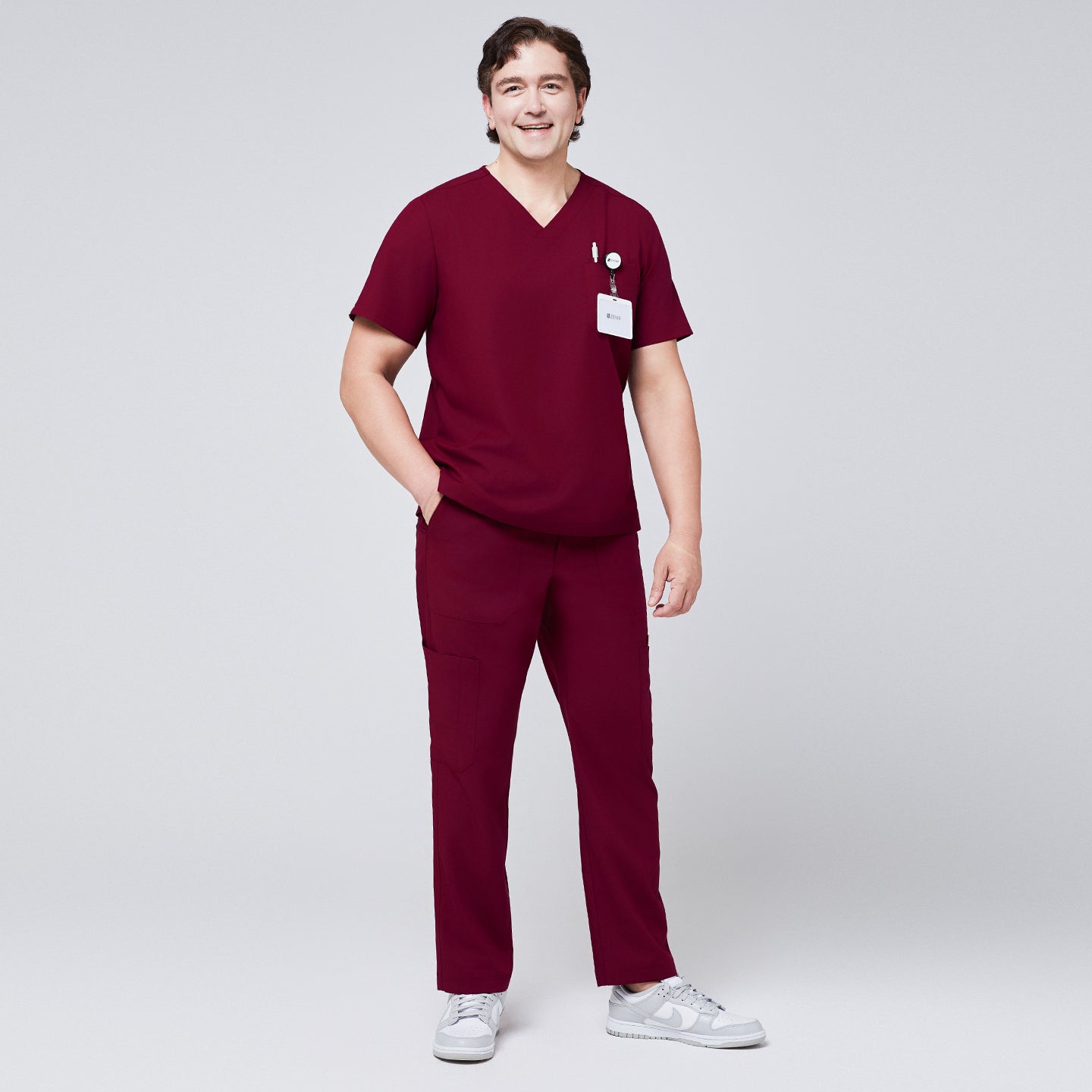 Man wearing burgundy V-neck scrub top and 8-pocket straight scrub pants, paired with white sneakers and a name badge,Burgundy