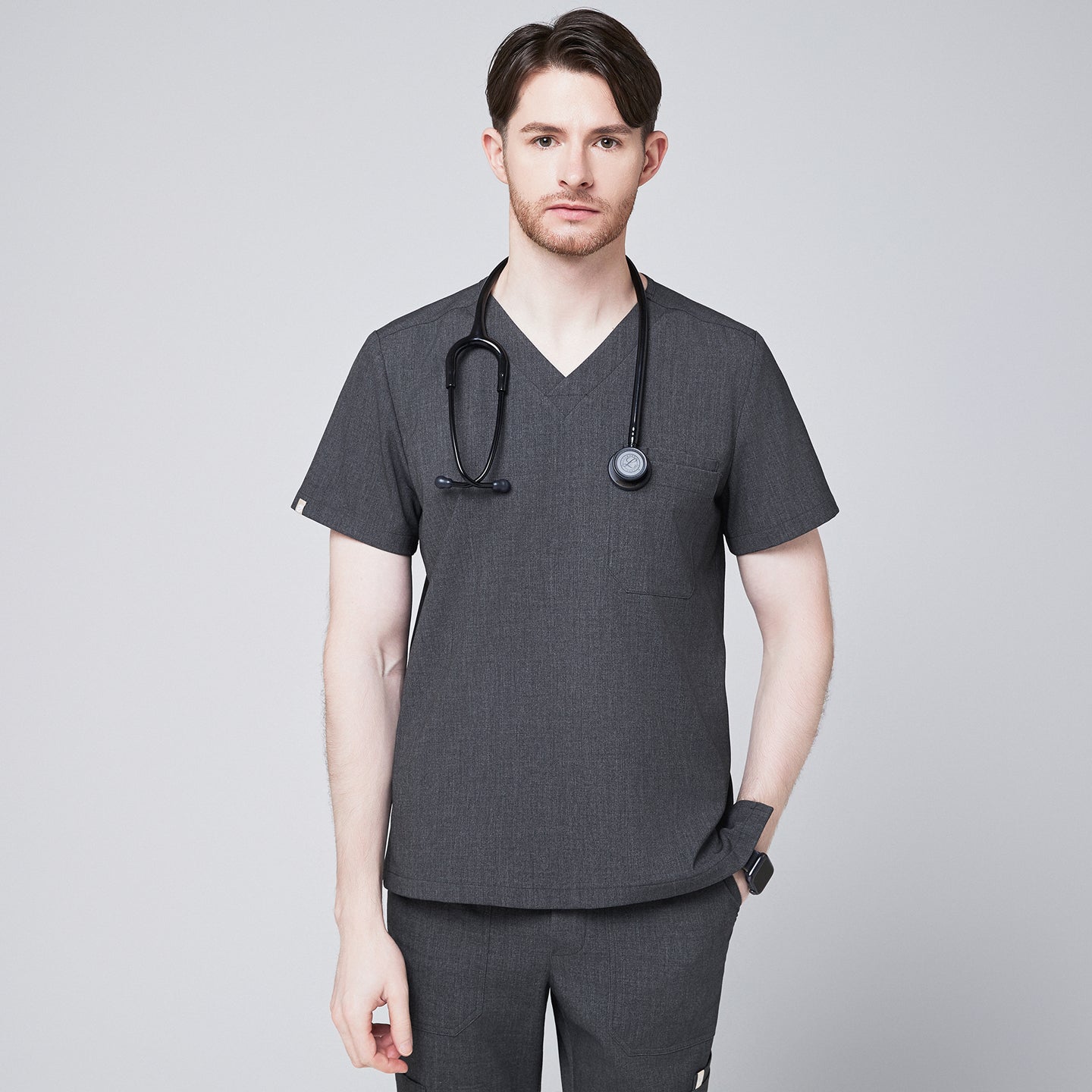 Male healthcare professional wearing a charcoal gray V-neck scrub top with three pockets and black stethoscope around neck, standing against a plain background,Charcoal Gray