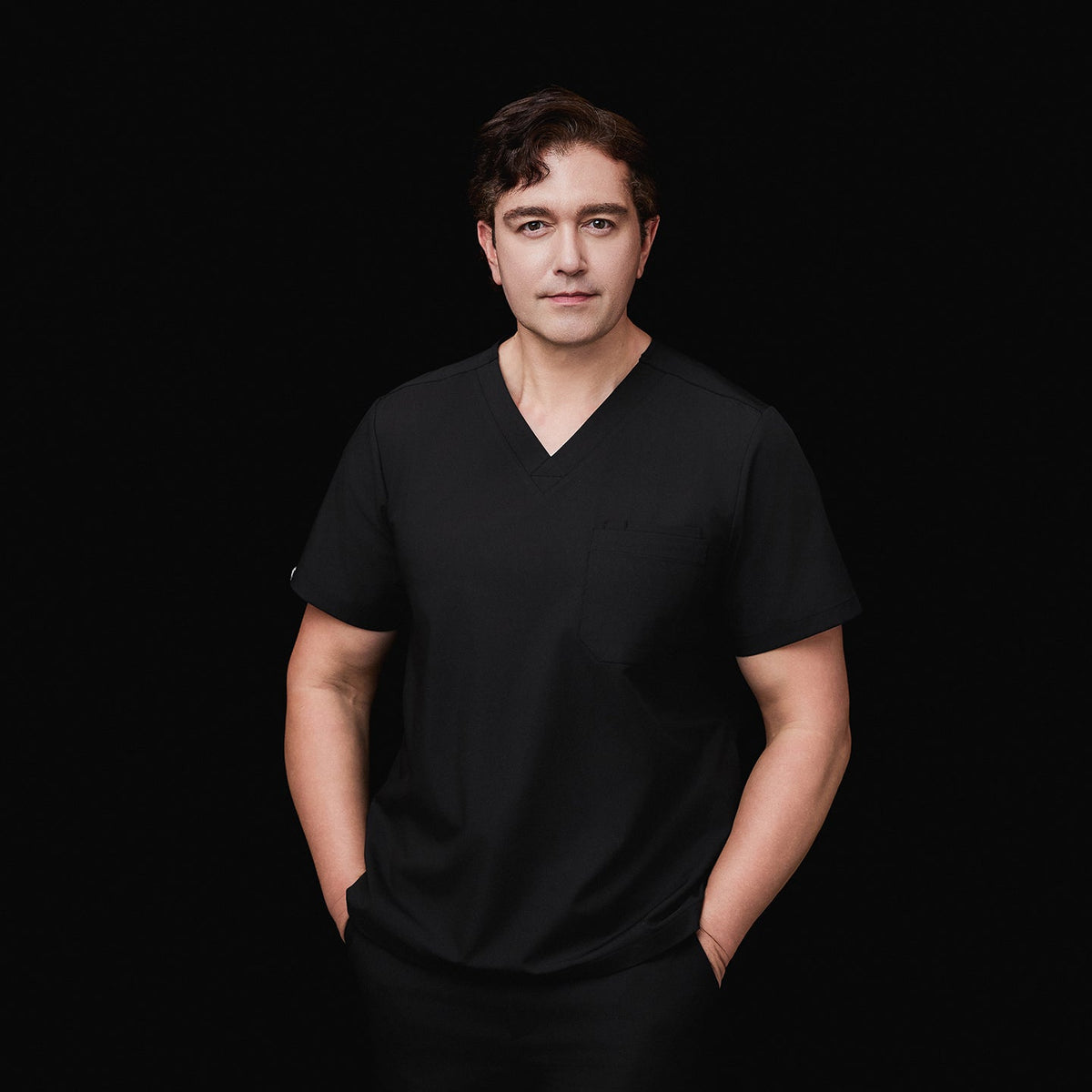 Model wearing an eco black 3-pocket scrub top with a V-neck design, hands in pockets, styled with matching scrub pants, against a black background,Eco Black