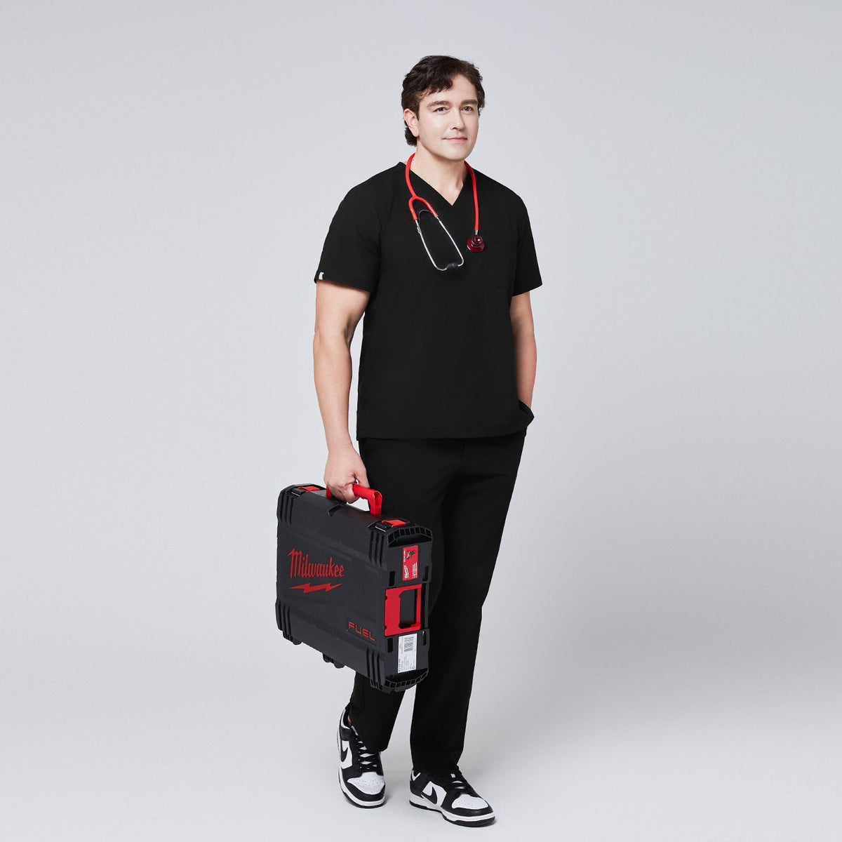 Full-body view of a model wearing an eco black 3-pocket scrub top and matching pants, styled with a red stethoscope and carrying a Milwaukee tool case, paired with black sneakers,Eco Black