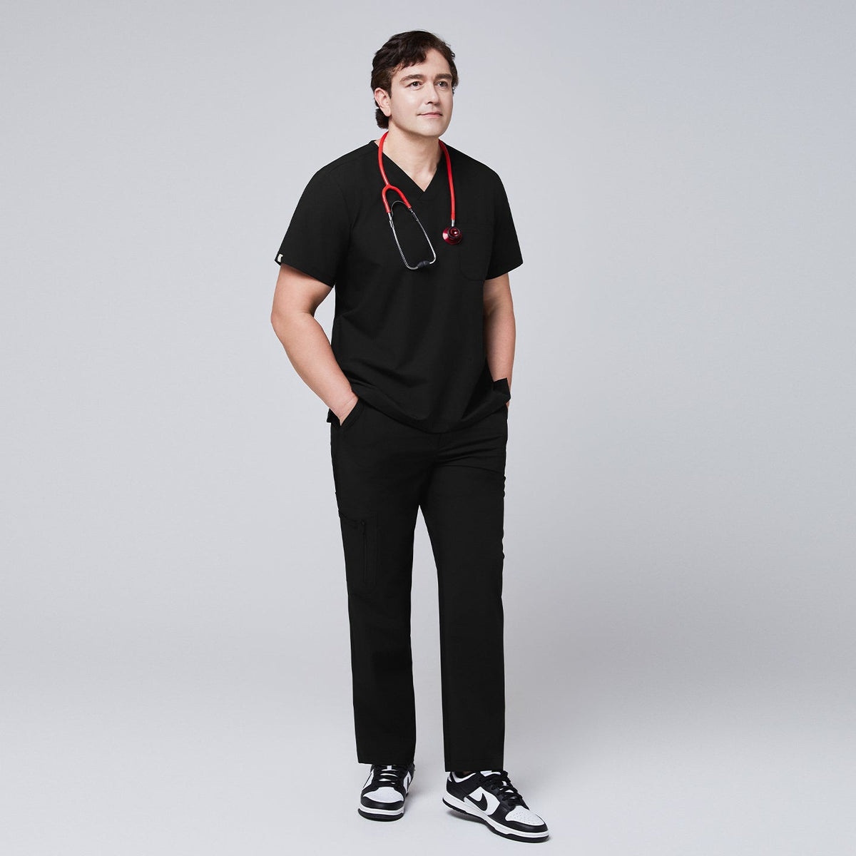 Full-body view of a model wearing an eco black 3-pocket scrub top and matching pants, styled with a red stethoscope and black sneakers,Eco Black