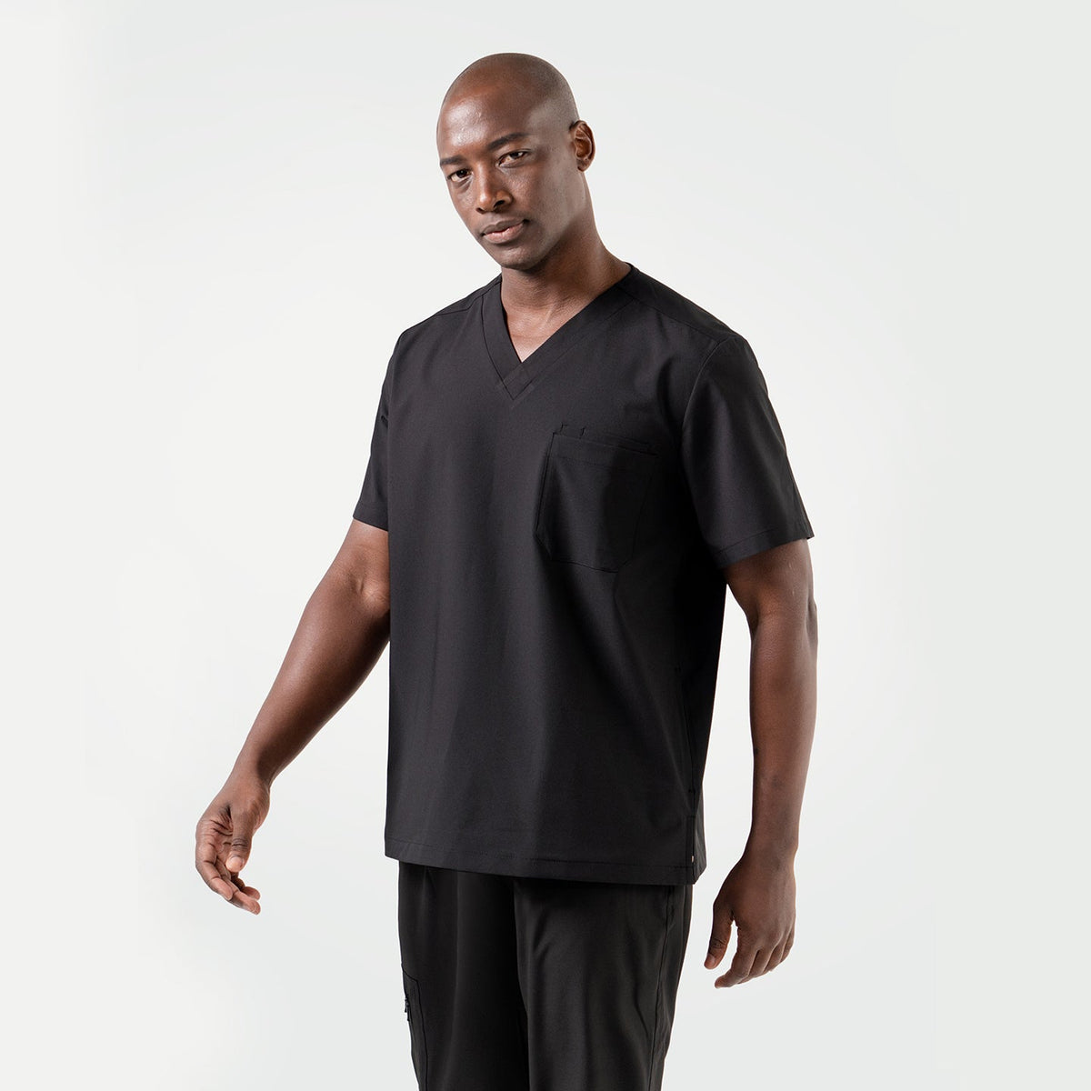 A smiling male model dressed in an Eco Black 3 Pocket scrub top, standing in a relaxed pose.