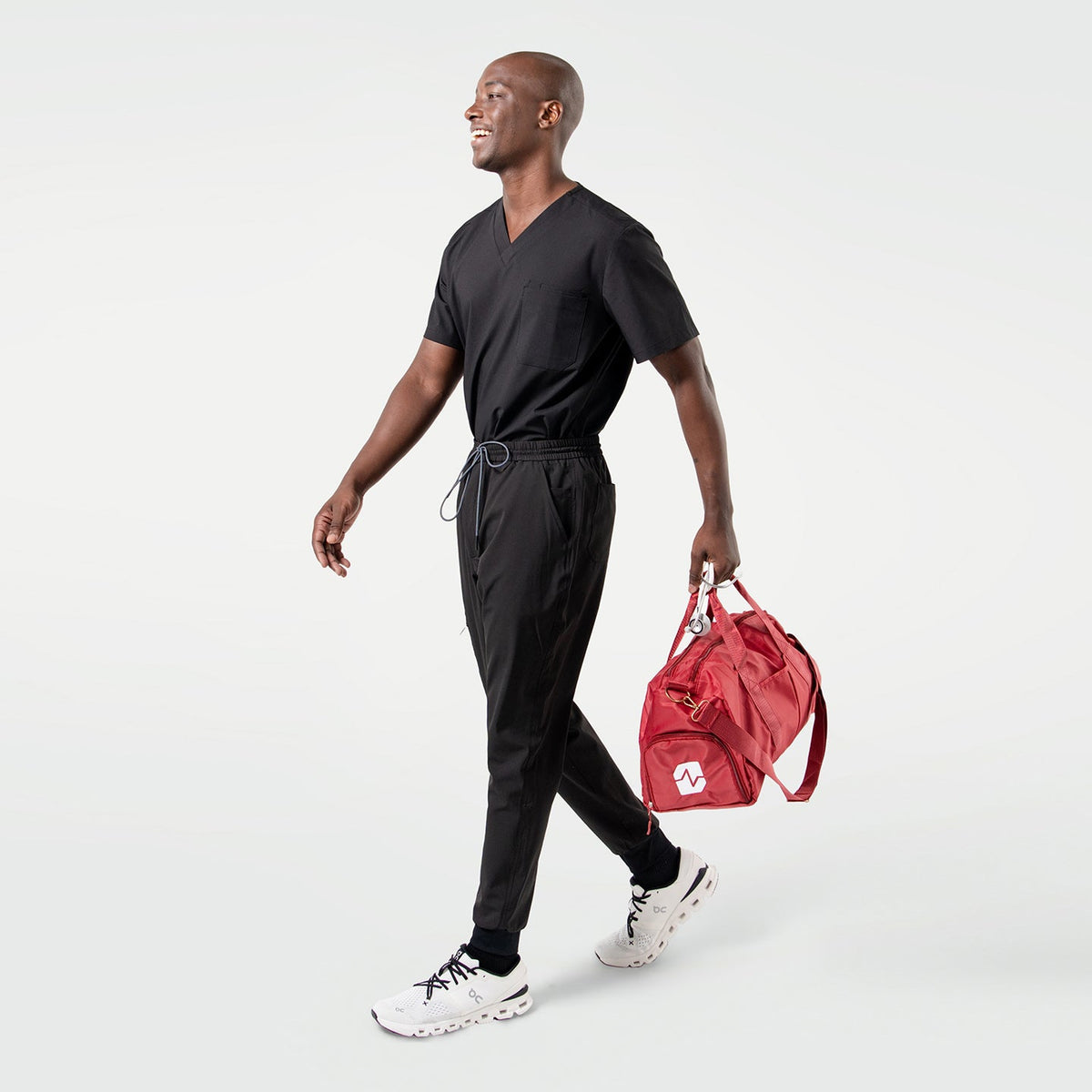 Smiling model in an Eco Black 3 pocket scrub top with short sleeves and chest pocket, paired with matching scrub pants while carrying a red duffel bag.