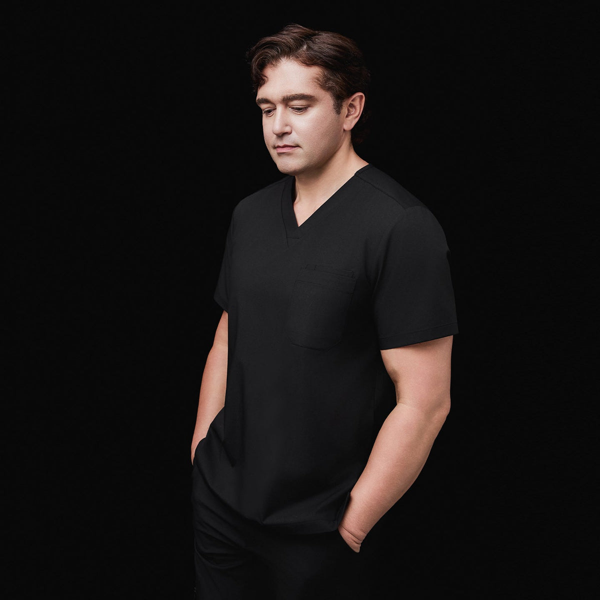 Side view of a model wearing an eco black 3-pocket scrub top with a V-neck design, hands in pockets, styled with matching scrub pants, against a black background,Eco Black