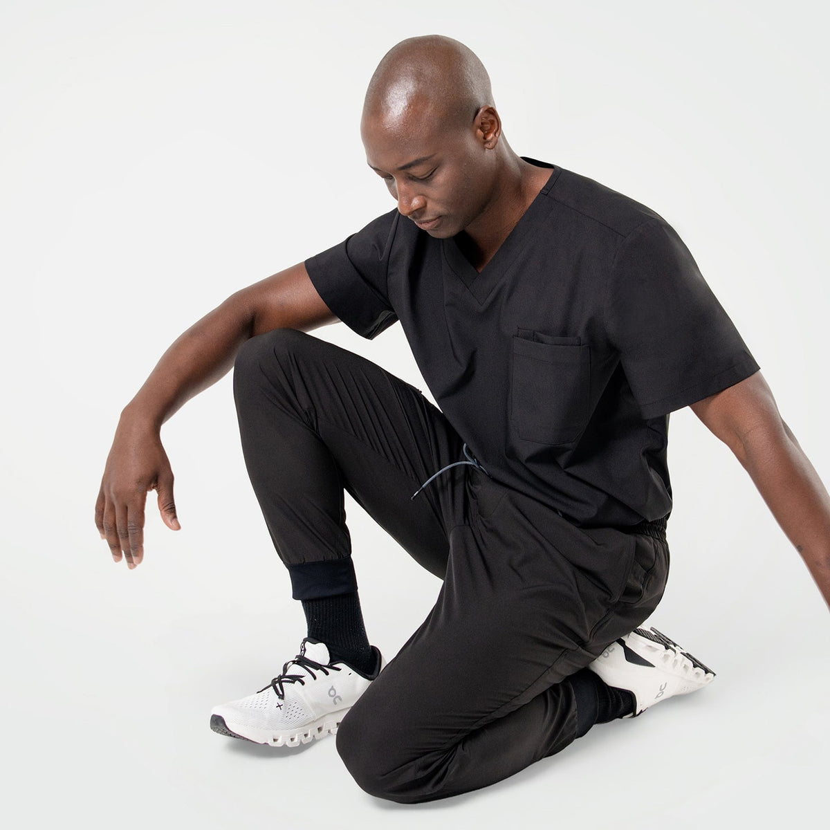 Kneeling pose in an Eco Black 3 pocket scrub top with short sleeves and chest pocket, paired with matching jogger scrub pants and white sneakers.