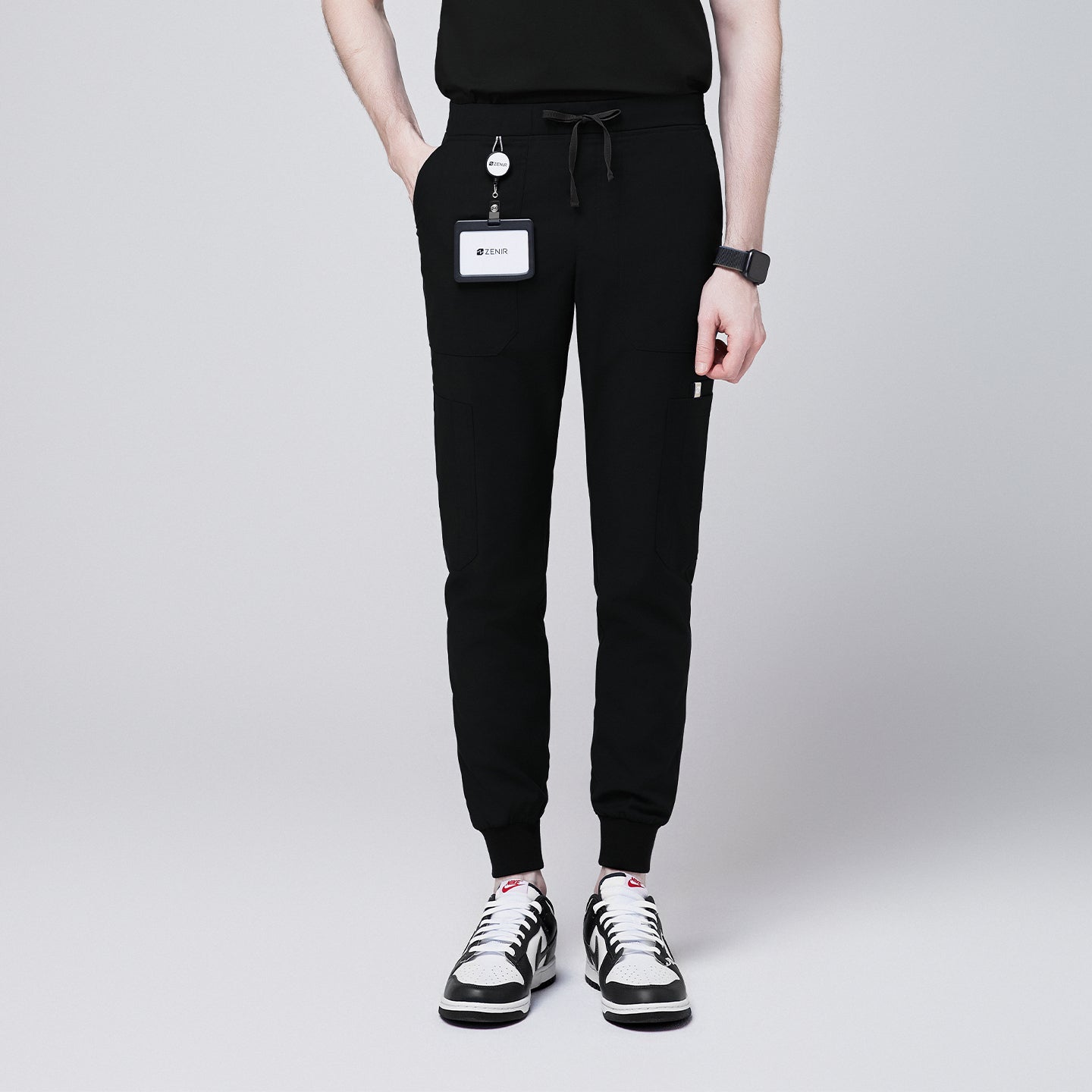 Front view of black jogger scrub pants with 8 pockets, drawstring waistband, and cuffed ankles, paired with black and white sneakers and a name badge,Eco Black