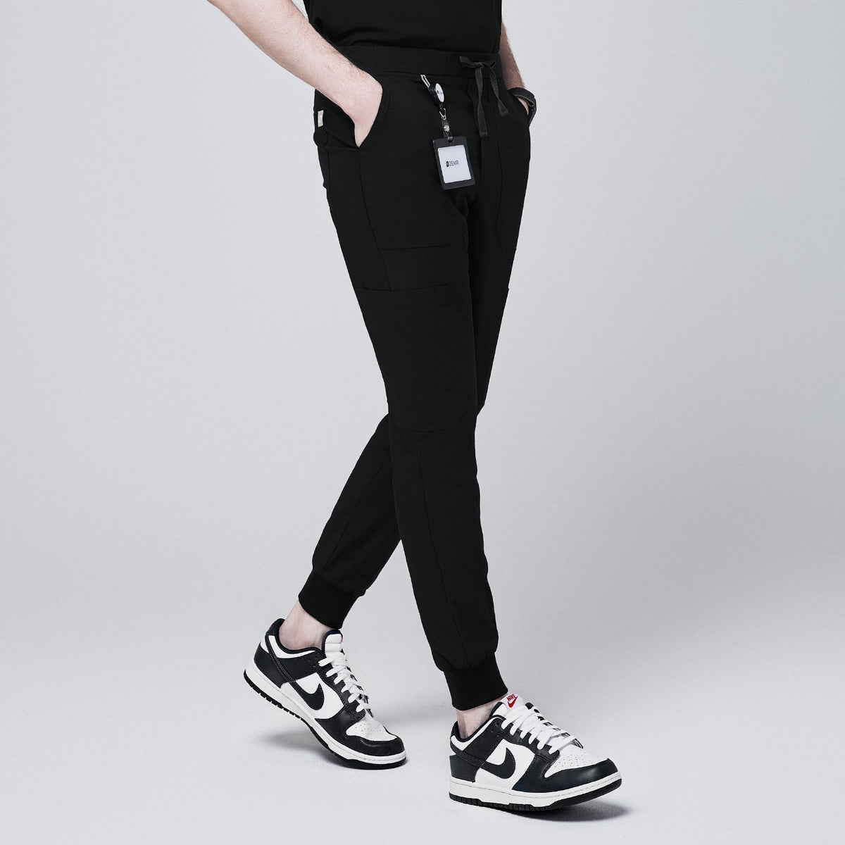 Side angle of black jogger scrub pants with 8 pockets, drawstring waistband, and cuffed ankles, paired with black and white sneakers and a name badge,Eco Black