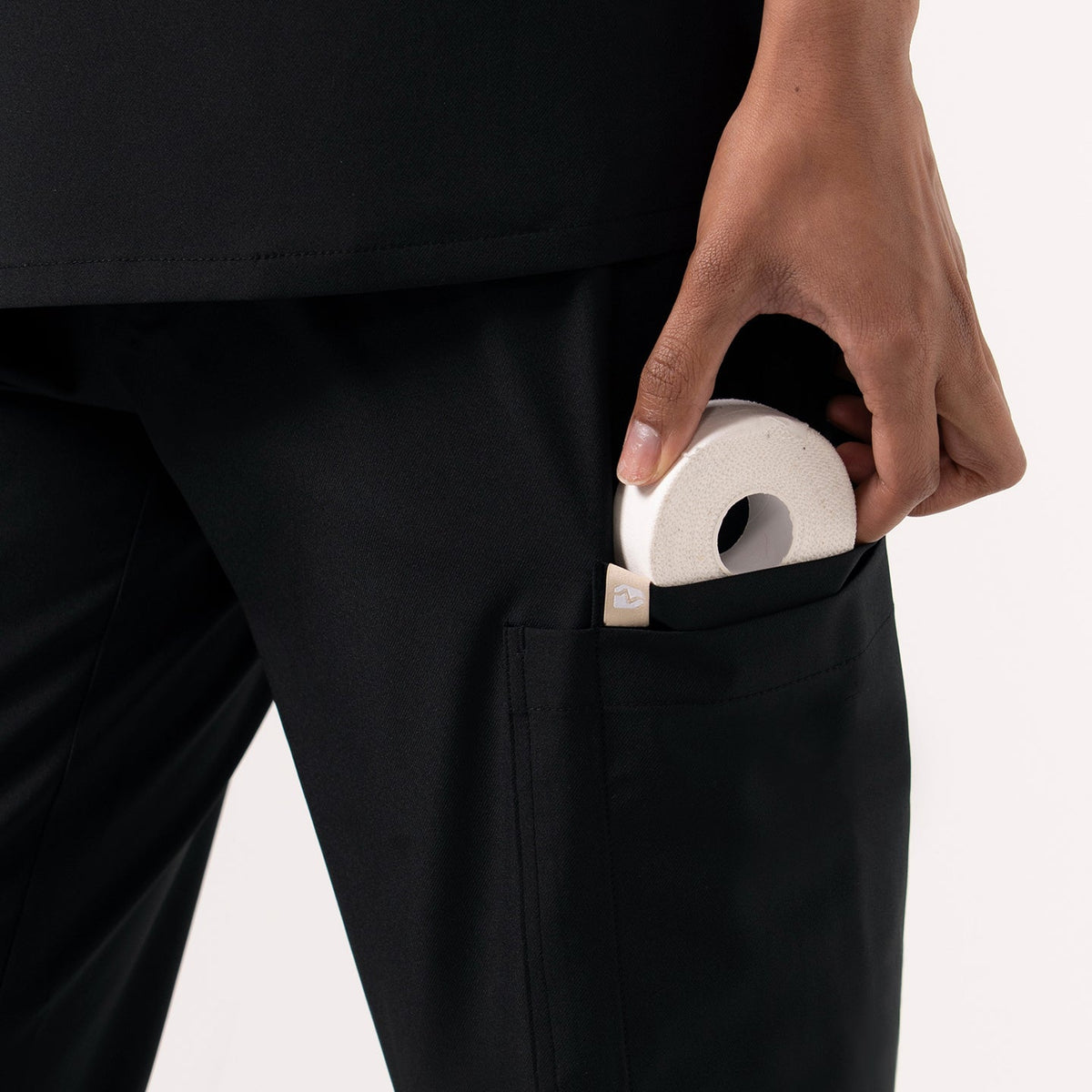 Close-up view of a person wearing black jogger-style scrub pants, shown placing a small round medical item into the side cargo pocket. The fabric appears smooth and durable, and the pocket design is deep and functional, emphasizing storage convenience for healthcare professionals.