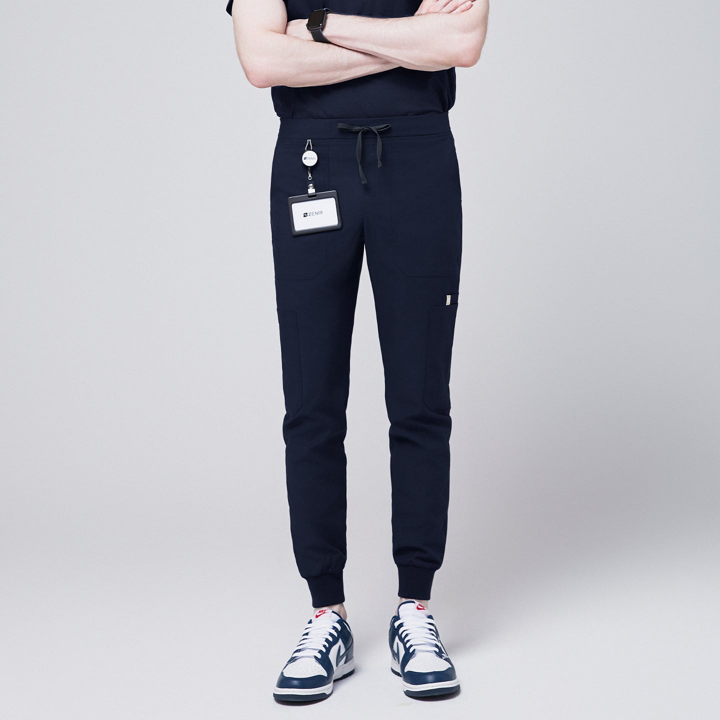 Front view of navy jogger scrub pants with 8 pockets, drawstring waistband, cuffed ankles, and a name badge, paired with navy sneakers,Eco Navy