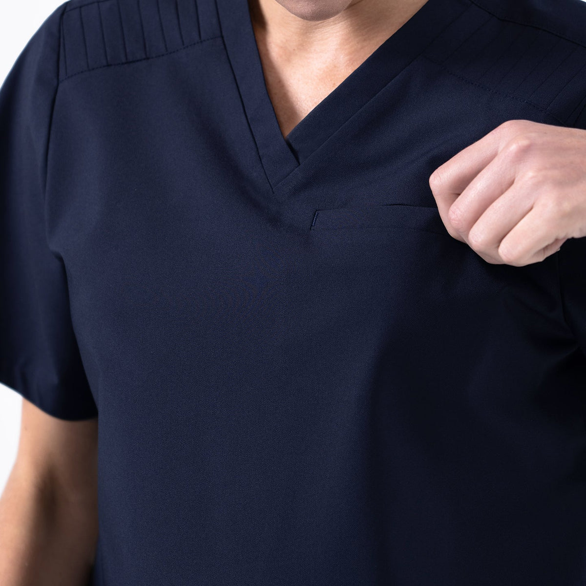 Close-up of the Eco-Navy pleated-shoulder scrub top, highlighting the stitched shoulder detailing and the subtle chest pocket as the model adjusts the fabric.