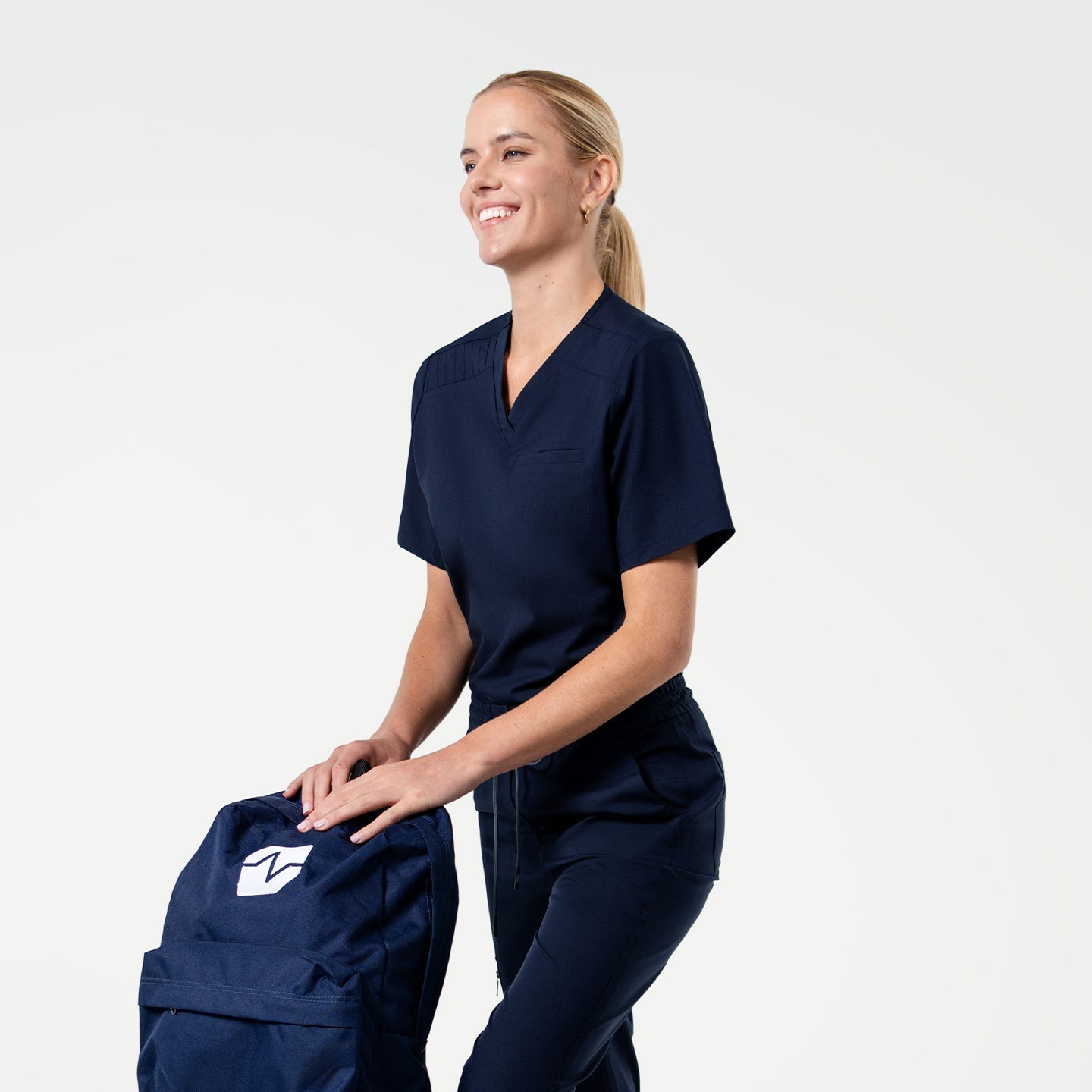 Smiling in the Women's Eco Navy Pleated Shoulder Scrub Top and pants, holding a navy backpack, upper body view