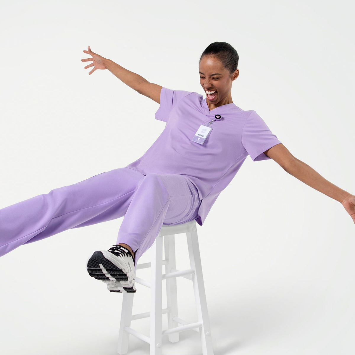 Energetic pose in the Women's Lavender Pleated Shoulder Scrub Top and pants, sitting on a stool with arms wide open, full body shot