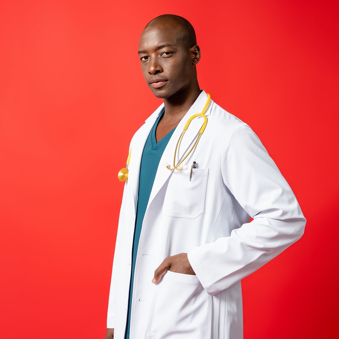 The LCM-02 Lab coat features a tailored fit with a chest pocket and lower patch pockets, styled with a yellow stethoscope draped around the neck. Model poses against a bold red background with one hand in the pocket.