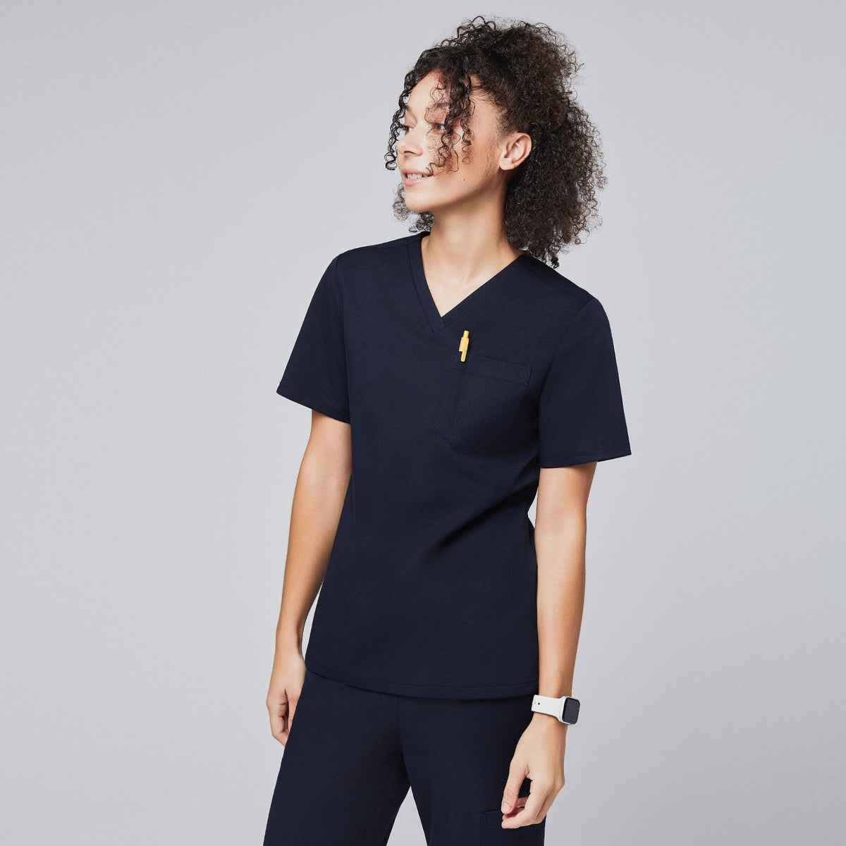 Woman wearing a navy scrub top with a chest pocket holding a yellow pen, looking to the side,Navy