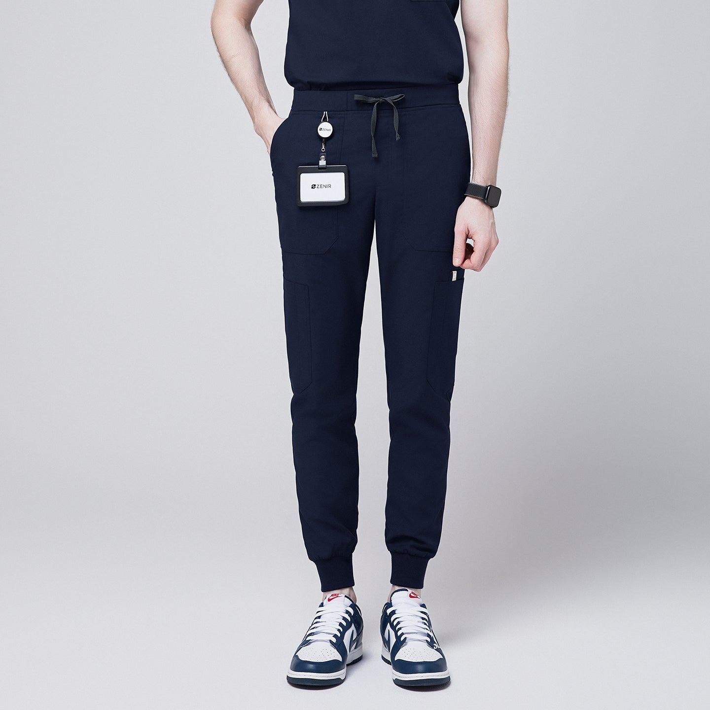 Front view of navy 8-pocket jogger scrub pants with drawstring waistband, cuffed ankles, and a name badge, paired with sneakers,Navy