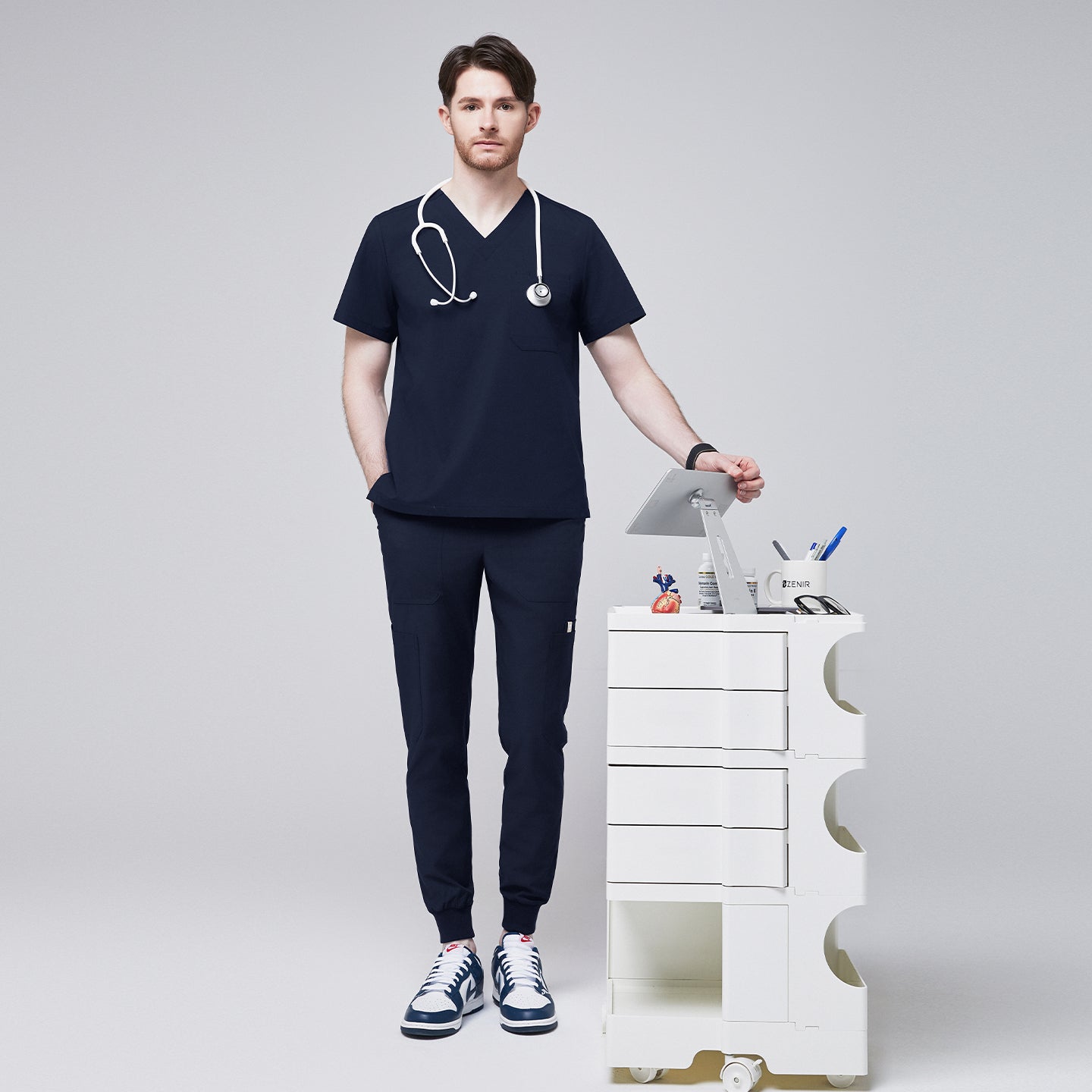 Man wearing navy V-neck scrub top and 8-pocket jogger scrub pants, accessorized with a stethoscope, standing beside a white medical cart,Navy