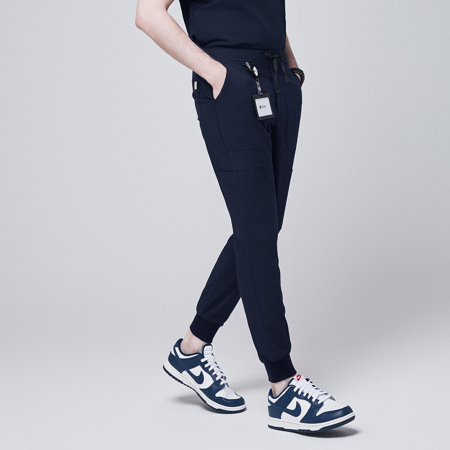 Side angle of navy jogger scrub pants with 8 pockets, drawstring waistband, and cuffed ankles, paired with navy sneakers and a name badge,Navy