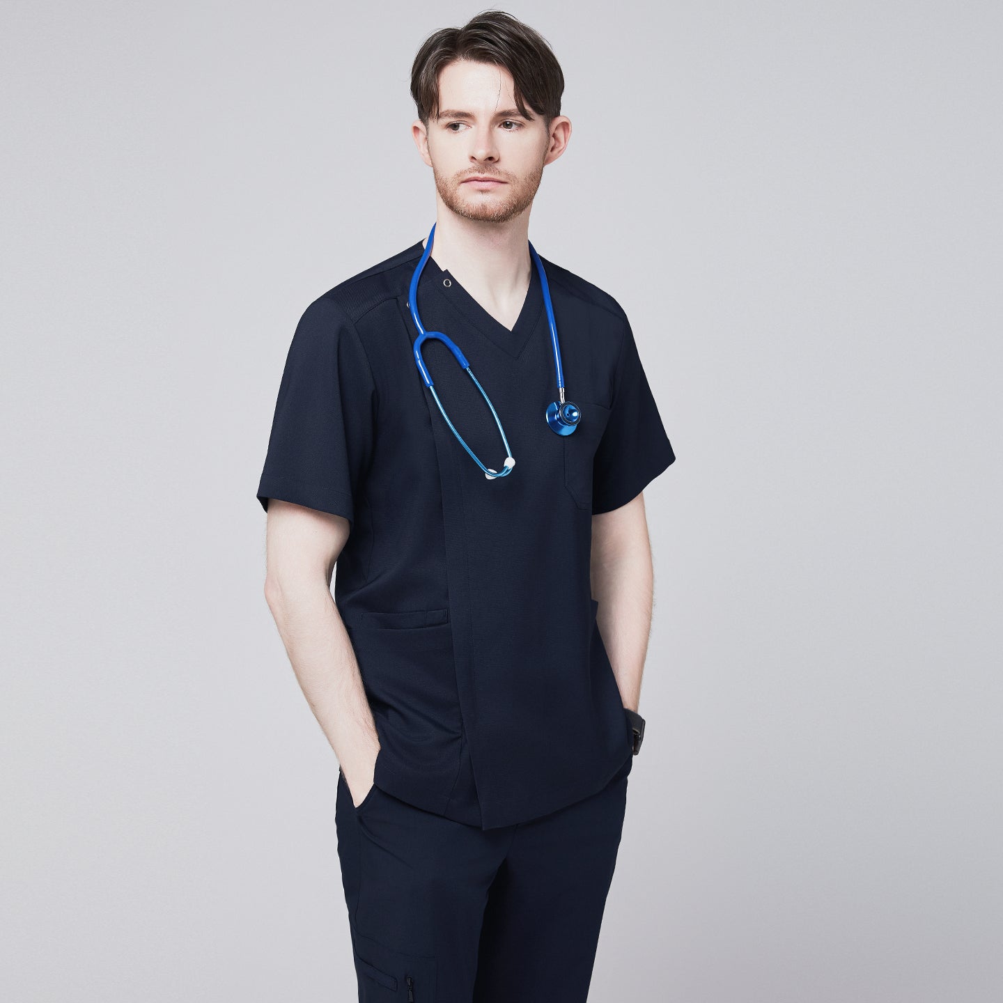Man wearing a navy scrub top with a front zipper detail, hands in pockets, styled with a stethoscope around his neck, looking confidently to the side,Navy