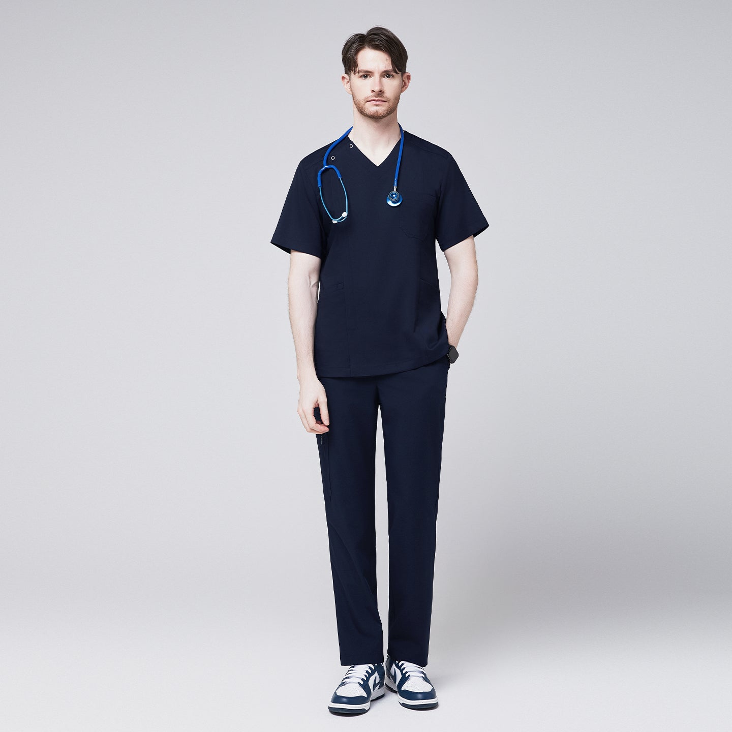 Full-body view of a man wearing a navy scrub top with a front zipper and matching pants, accessorized with a blue stethoscope and casual sneakers,Navy