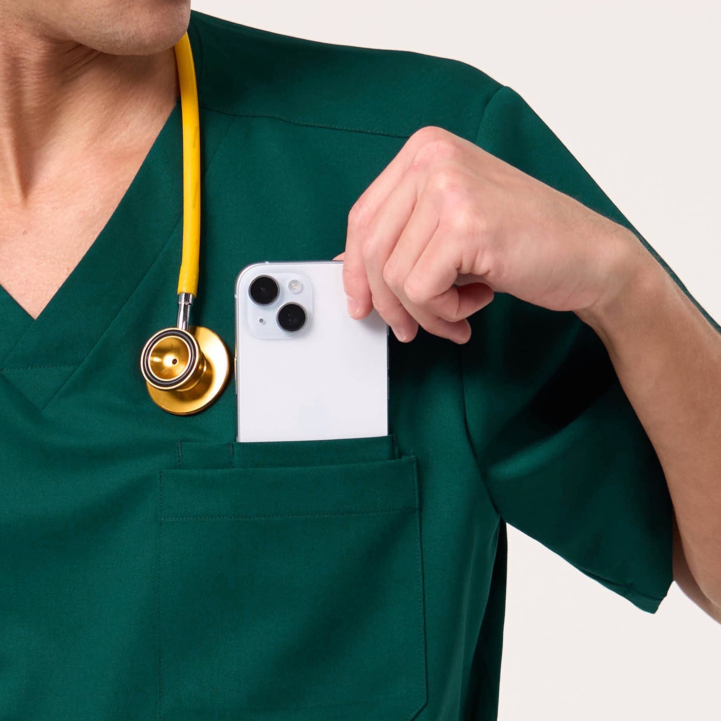 Wearing a Pine Green scrub top, the male model places a smartphone into the chest pocket while a yellow stethoscope hangs around his neck.