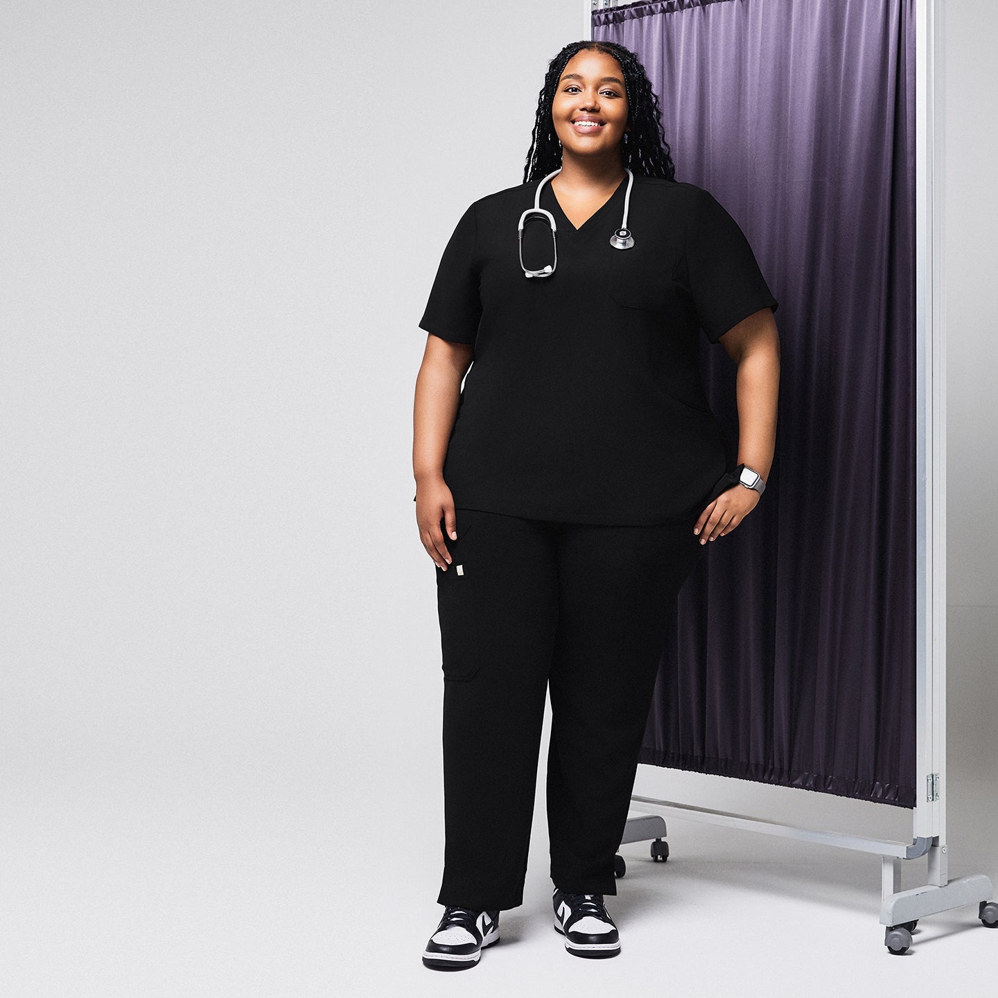 Female medical professional in a black scrub set with a stethoscope around her neck, standing next to a privacy curtain, smiling with her hand in her pocket,Rich Black
