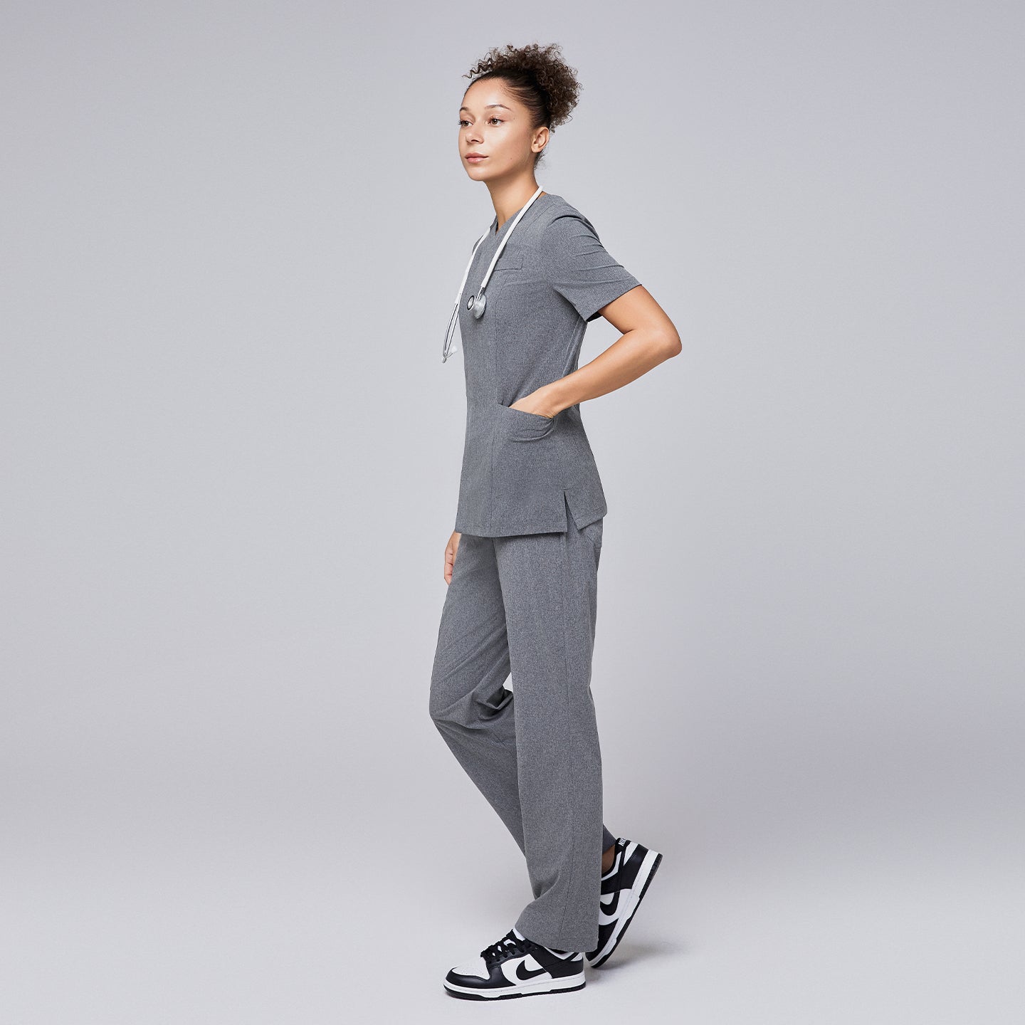 Woman wearing Zenir soft gray front zipper scrub set with hands in pockets, paired with a stethoscope and black and white sneakers,Ash Gray