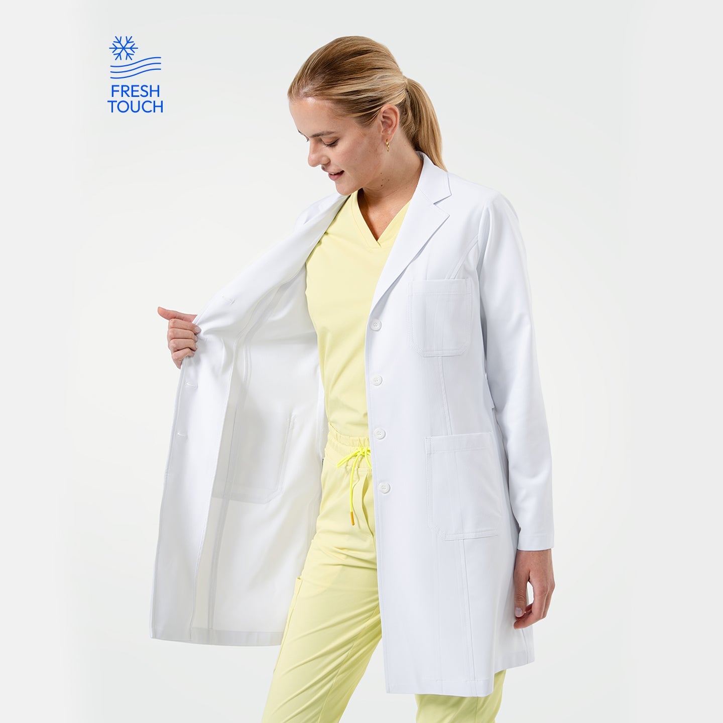 Female doctor wearing a white lab coat over yellow scrubs, looking down while opening the coat to show the inner lining on a light gray background with a “Fresh Touch” logo.