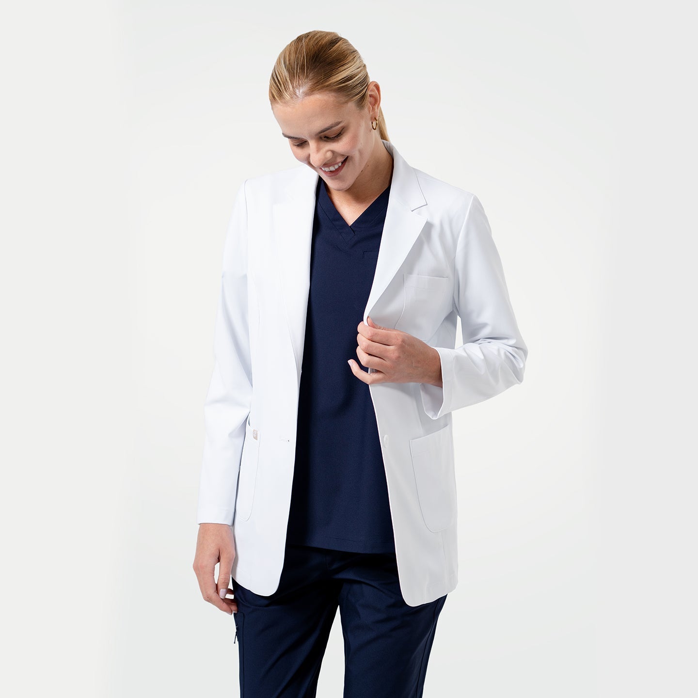 Smiling female healthcare professional wearing a white lab coat over navy scrubs, looking down with a gentle expression on a light gray background.