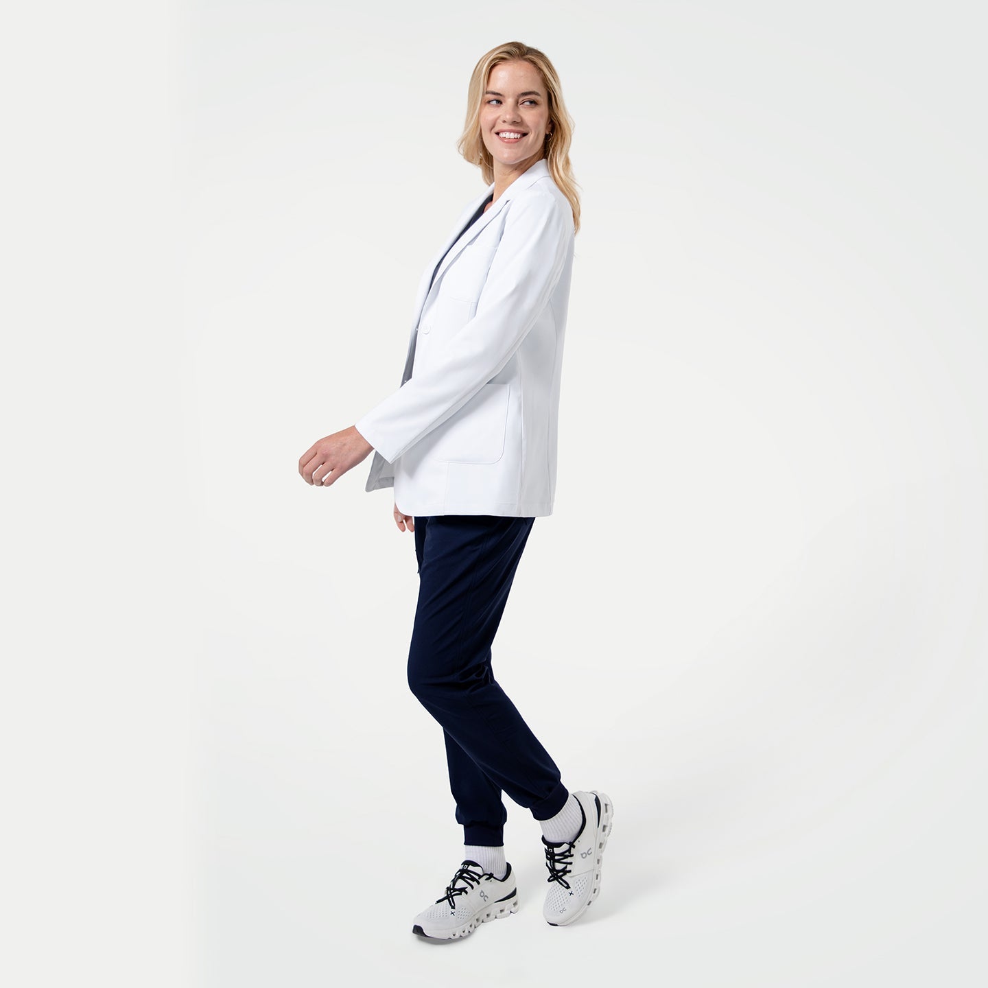 Woman wearing a white lab coat, navy pants, and white sneakers on a white background
