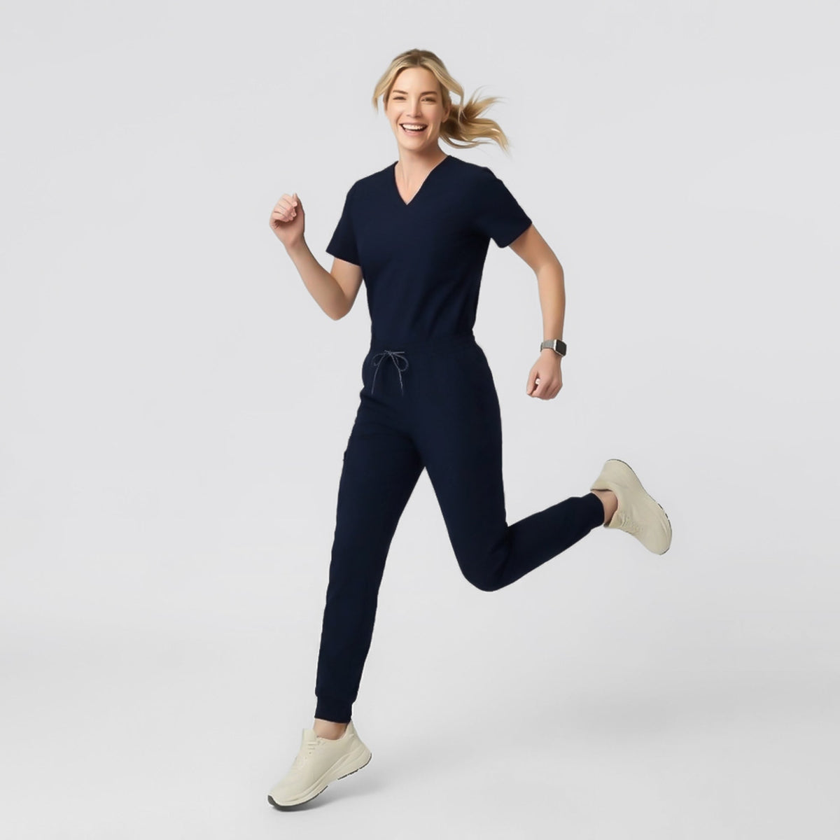 Model walking in a navy V-neck pleated-shoulder scrub top and matching jogger scrub pants, hands in pockets, against a clean light background.