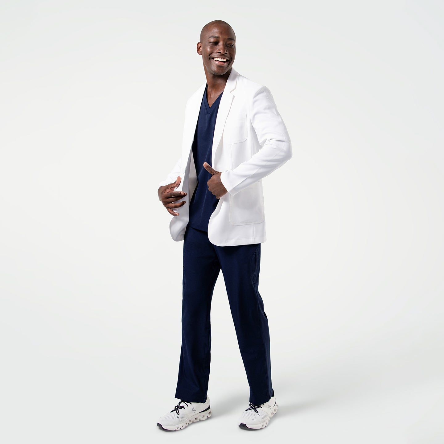 Male model in the Men's LCM-100 Lab Coat in White over medical scrubs, full body view, smiling while standing with hands near the coat pockets