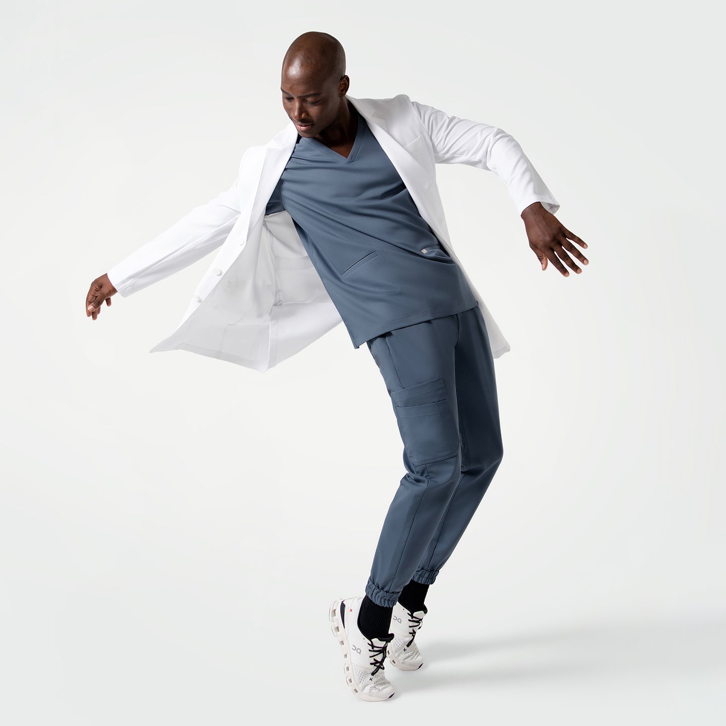 Male model in the Men's LCM-101 Lab Coat in White over medical scrubs, full body view, captured mid-motion with arms extended and body slightly leaning forward
