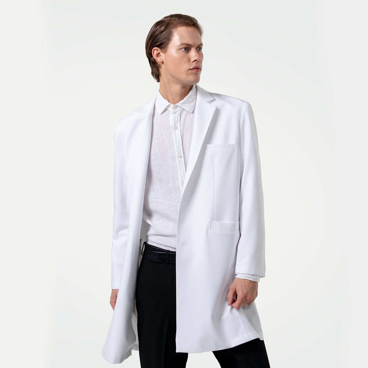 Looking to the side, a male doctor wears a white lab coat over a white dress shirt and black pants on a clean light gray background.