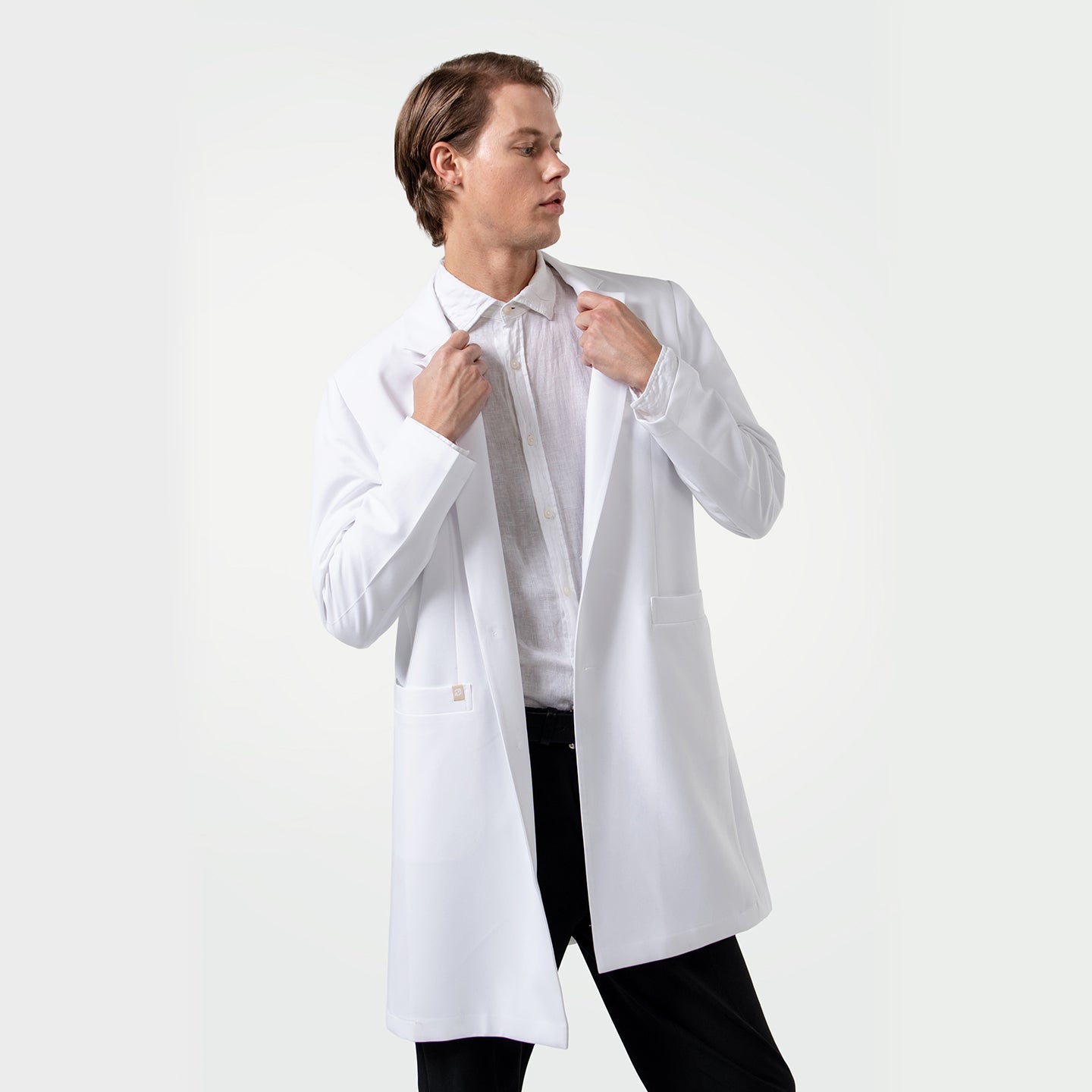 Confident male healthcare professional wearing a white coat over a white dress shirt, adjusting the lapels of the coat while looking to the side on a clean white background.