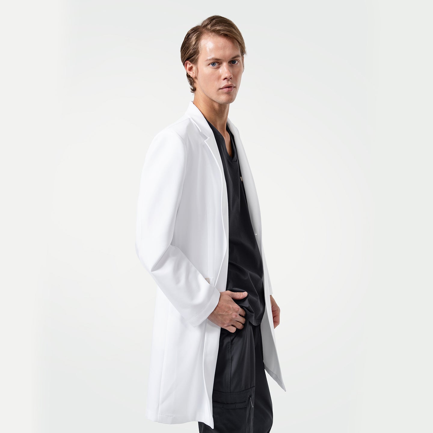 Lcm-103 white lab coat for men, shown on model wearing black scrubs underneath, standing sideways with one hand in pocket against light background.